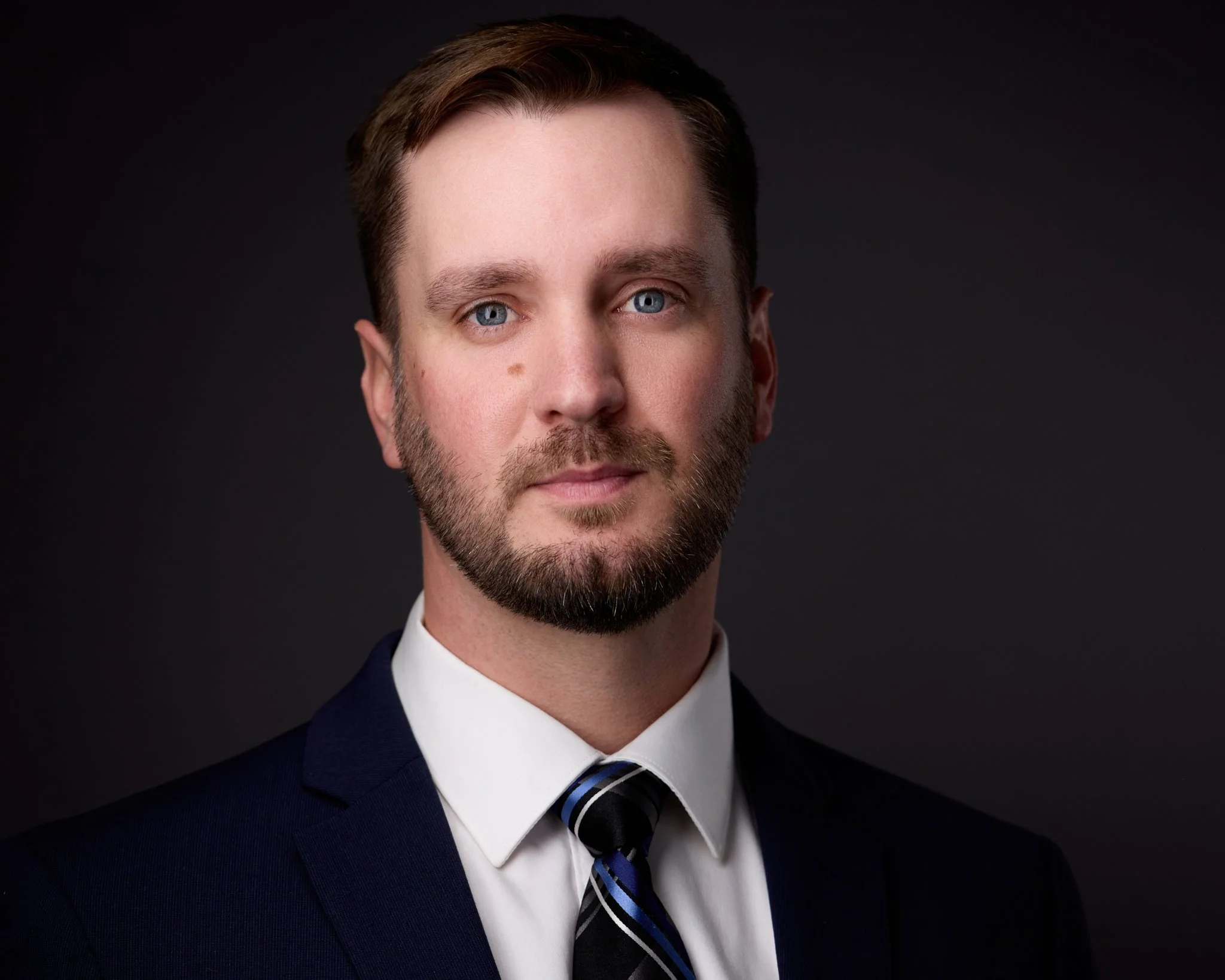 Portrait of a man with short brown hair, blue eyes, and a beard, wearing a dark suit, white shirt, and striped tie, against a black background.