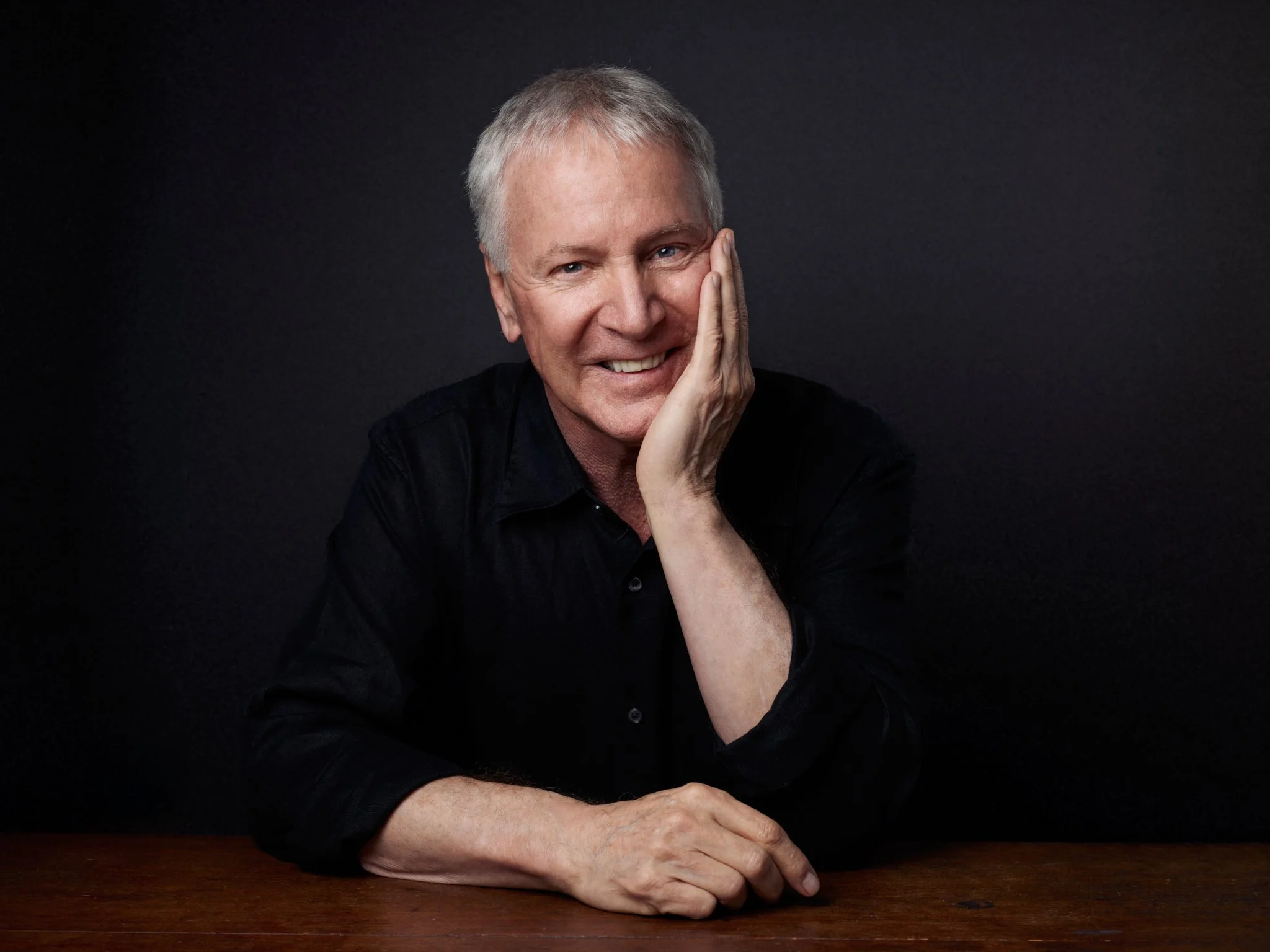 Portrait of a smiling older man with gray hair, resting his face on his hand, seated at a wooden table against a dark background.