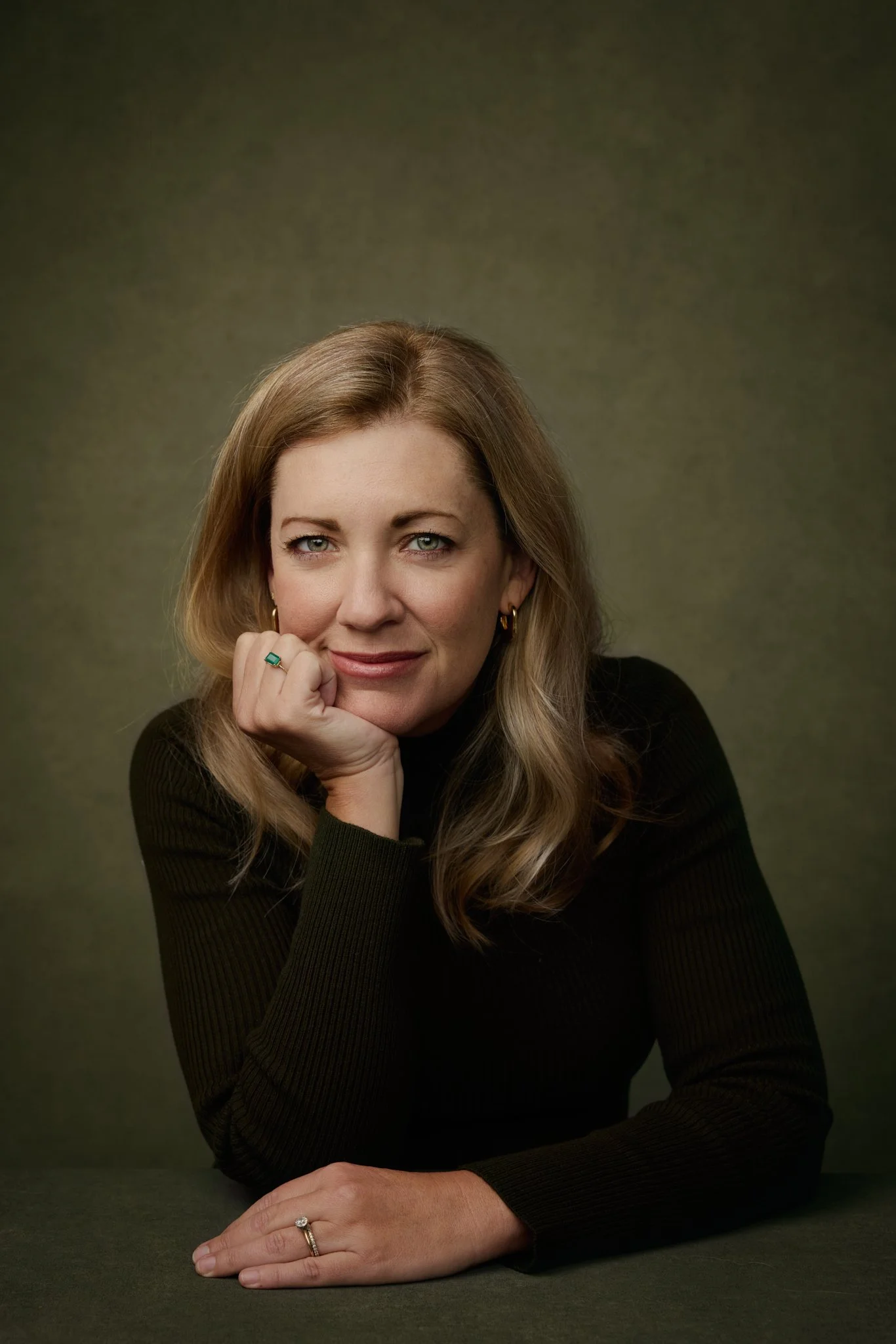 Portrait of a woman with blonde hair wearing a black top and jewelry, resting her chin on her hand, looking at the camera with a slight smile.