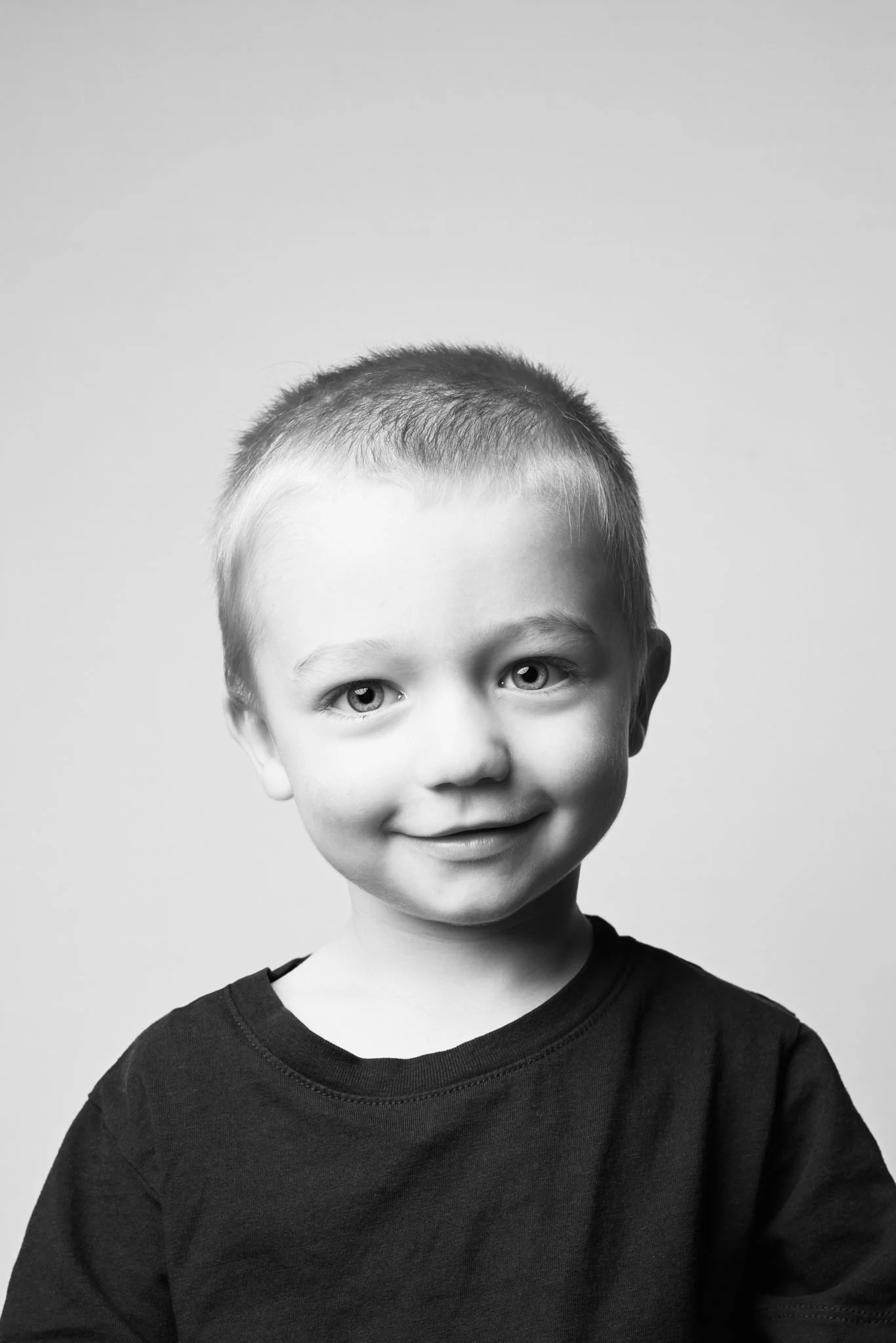 Black and white portrait of a young boy with short hair, smiling, wearing a dark T-shirt, against a plain light background.