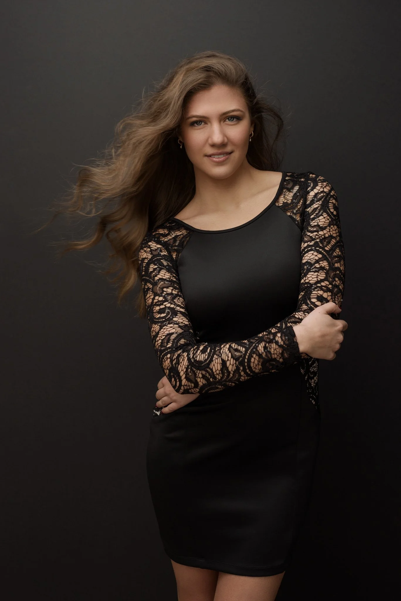 A young woman with long, wavy brown hair looks at the camera, wearing a black dress with lace long sleeves, standing against a dark background.
