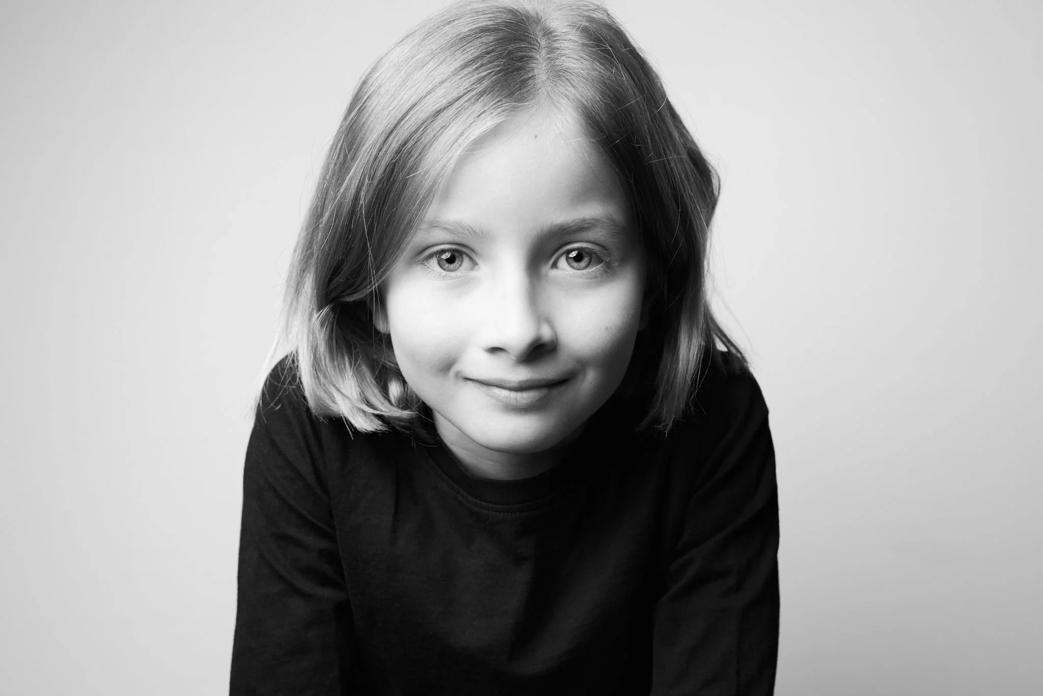 Black and white portrait of a young girl with shoulder-length hair, wearing a dark top, smiling gently at the camera with a neutral background.