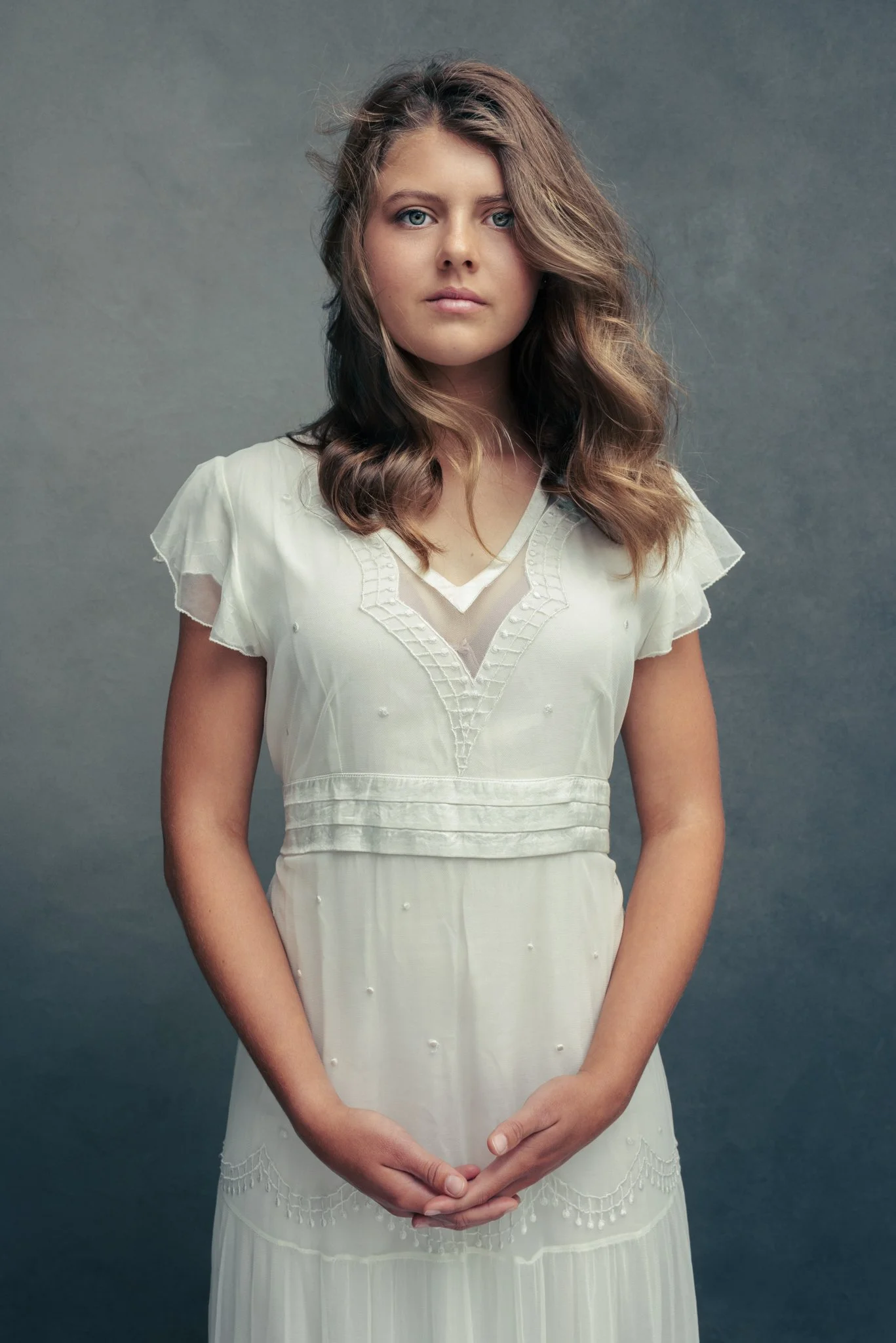 A young woman with wavy brown hair and blue eyes stands against a gray background, wearing a vintage white dress with embroidery and sheer details.