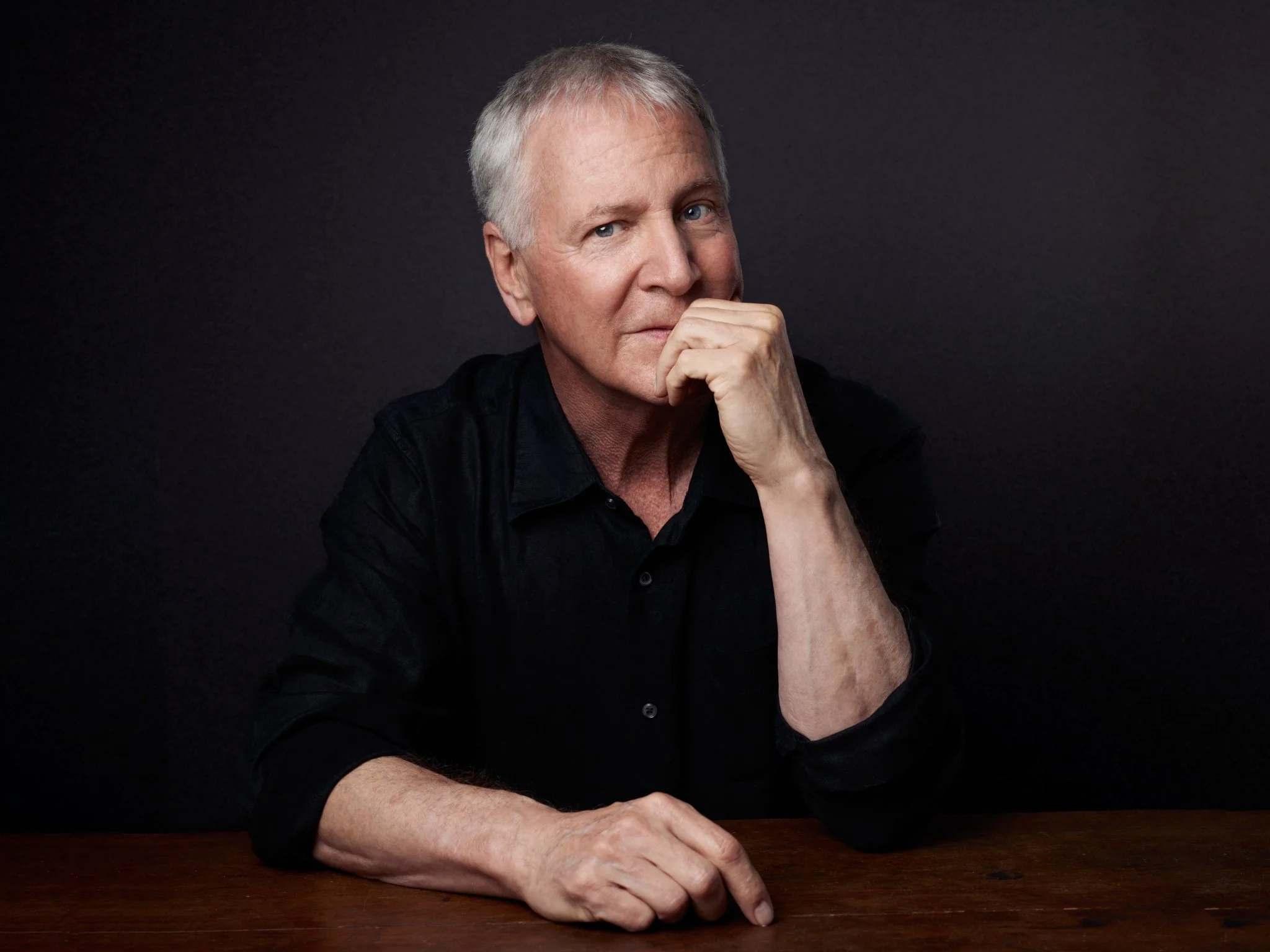 Portrait of an elderly man with gray hair, wearing a black shirt, sitting at a wooden table against a dark background. He has his right hand resting on the table and his left hand near his face, with a thoughtful expression.