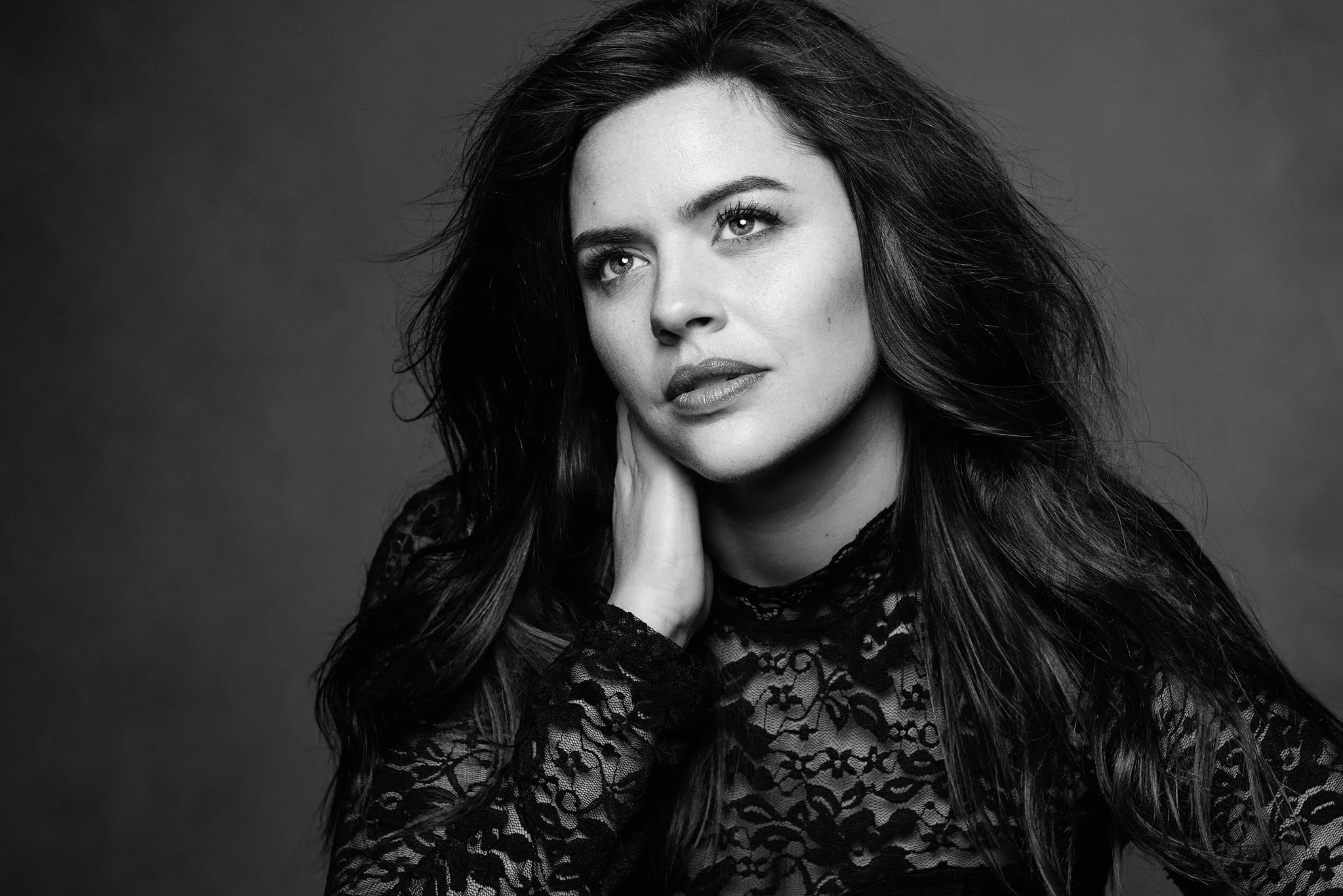 Black and white portrait of a woman with long, dark hair, wearing a lace top, looking directly at the camera with a soft expression, against a plain background.