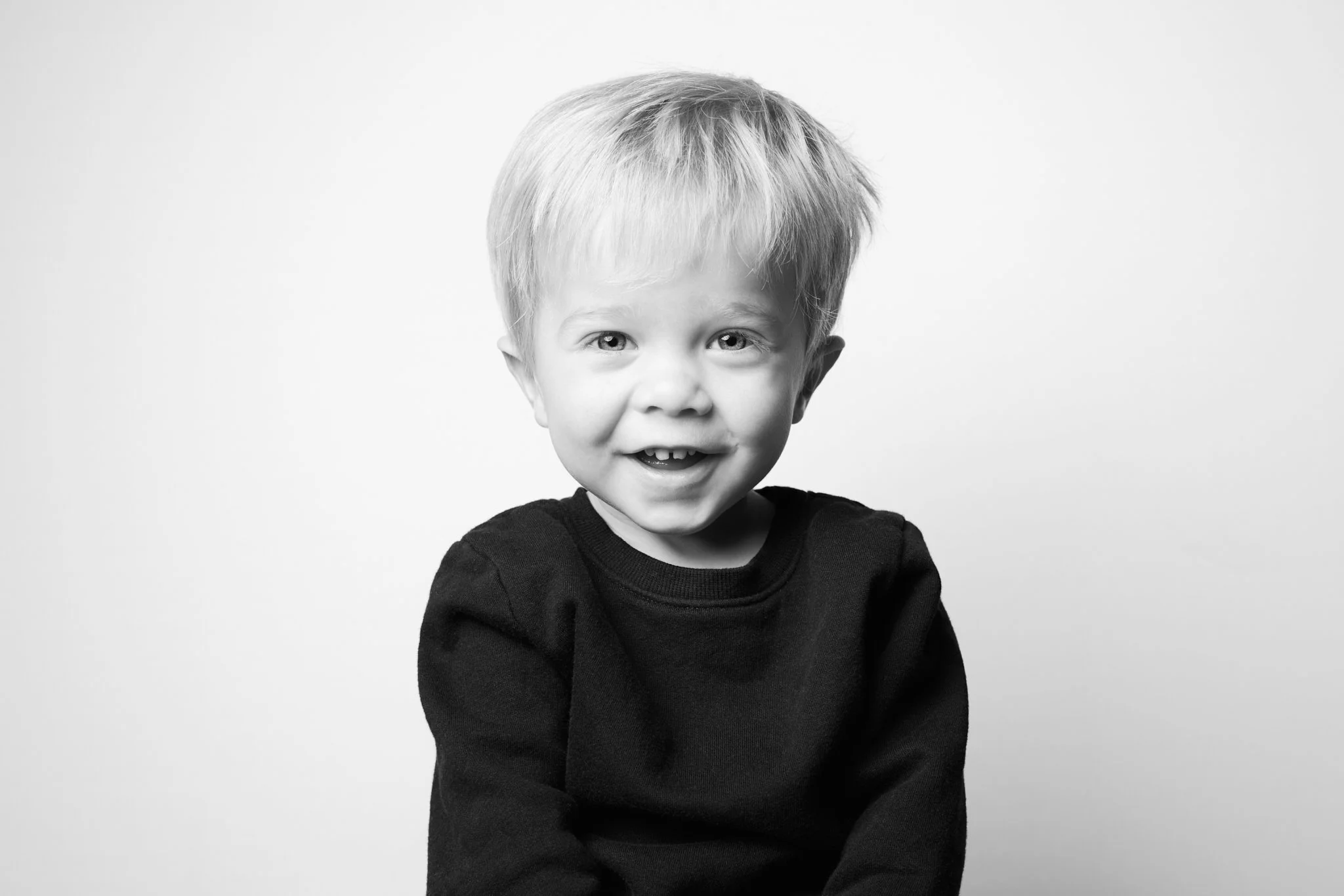 Black and white photo of a young boy with blonde hair, smiling, wearing a black long-sleeve shirt, against a plain white background.