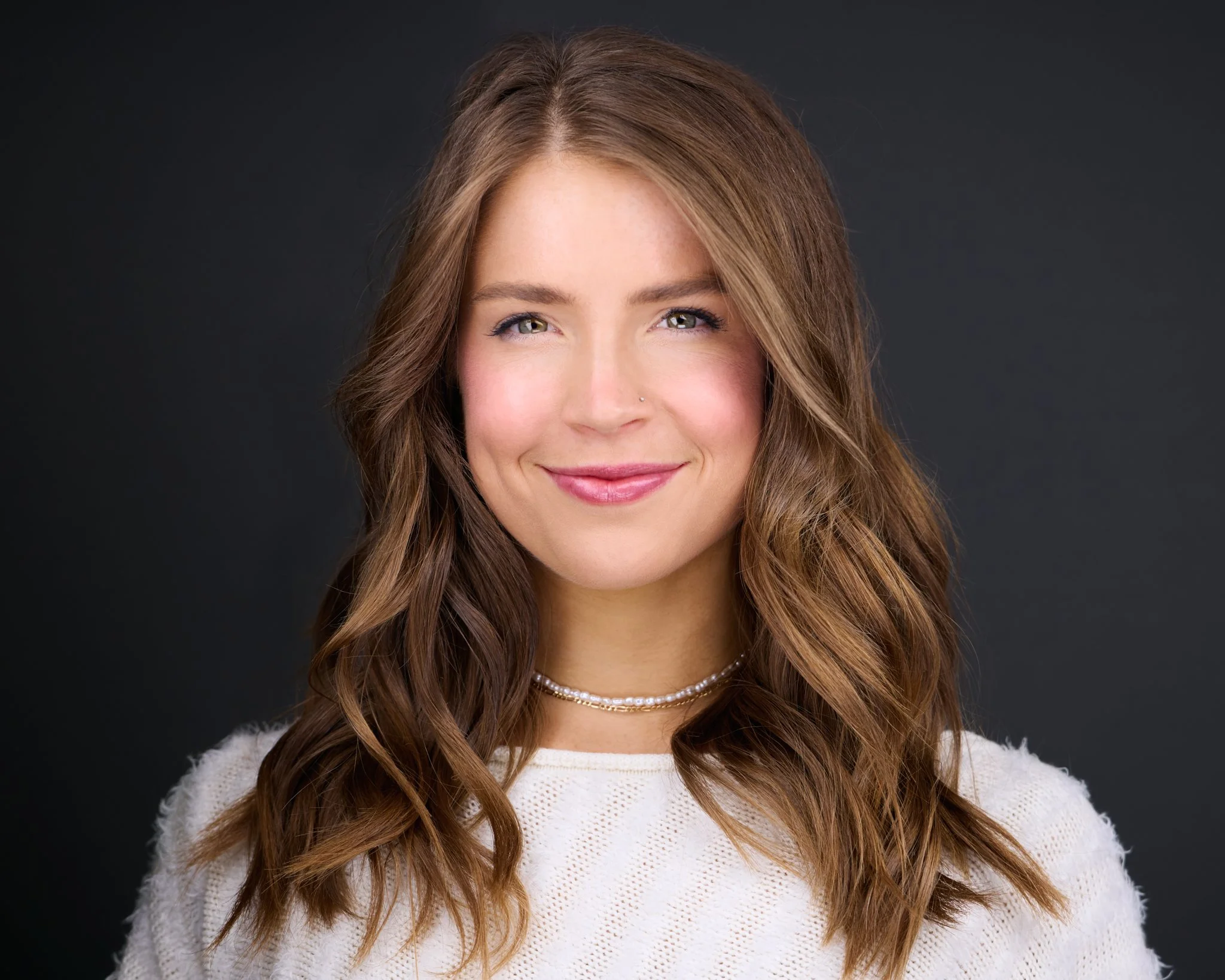 A smiling woman with long, wavy brown hair, wearing a white sweater and a layered pearl necklace against a dark background.