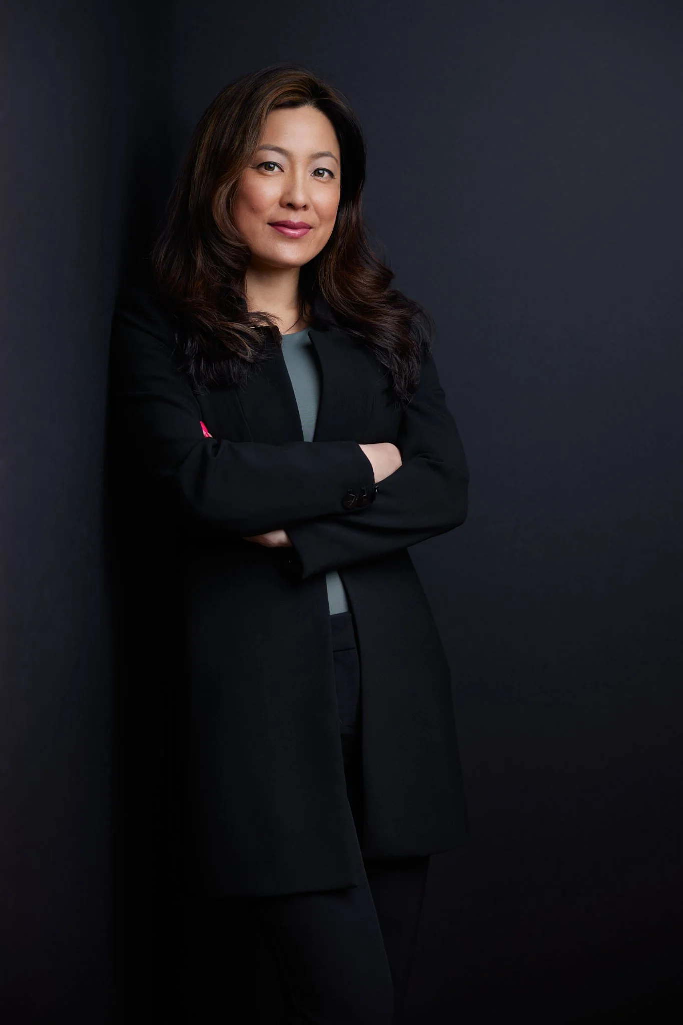 Portrait of a woman with long dark hair, wearing a black blazer and gray top, standing against a dark background.