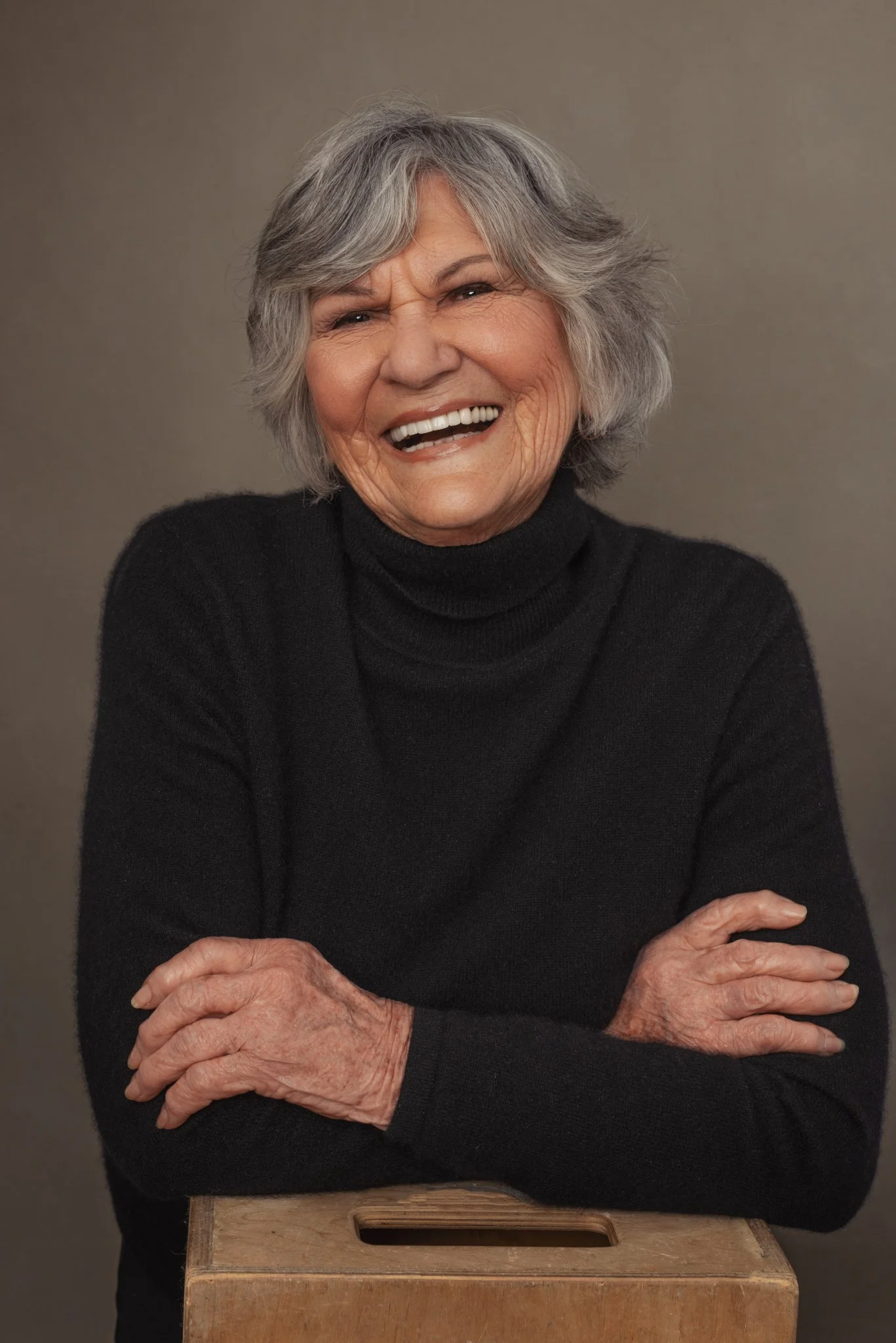 Smiling elderly woman with gray hair, wearing a black turtleneck, crossed arms, standing behind a wooden box, against a plain background.