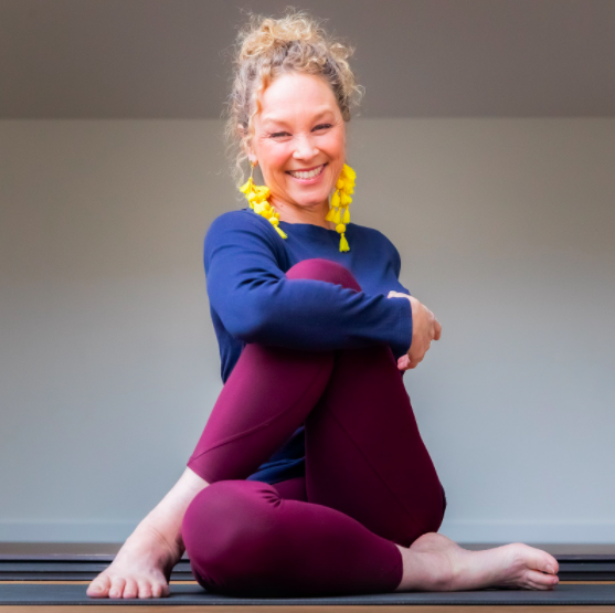 Woman sitting cross-legged on the floor, smiling, wearing a blue top, maroon leggings, and yellow earrings, in a brightly lit room.