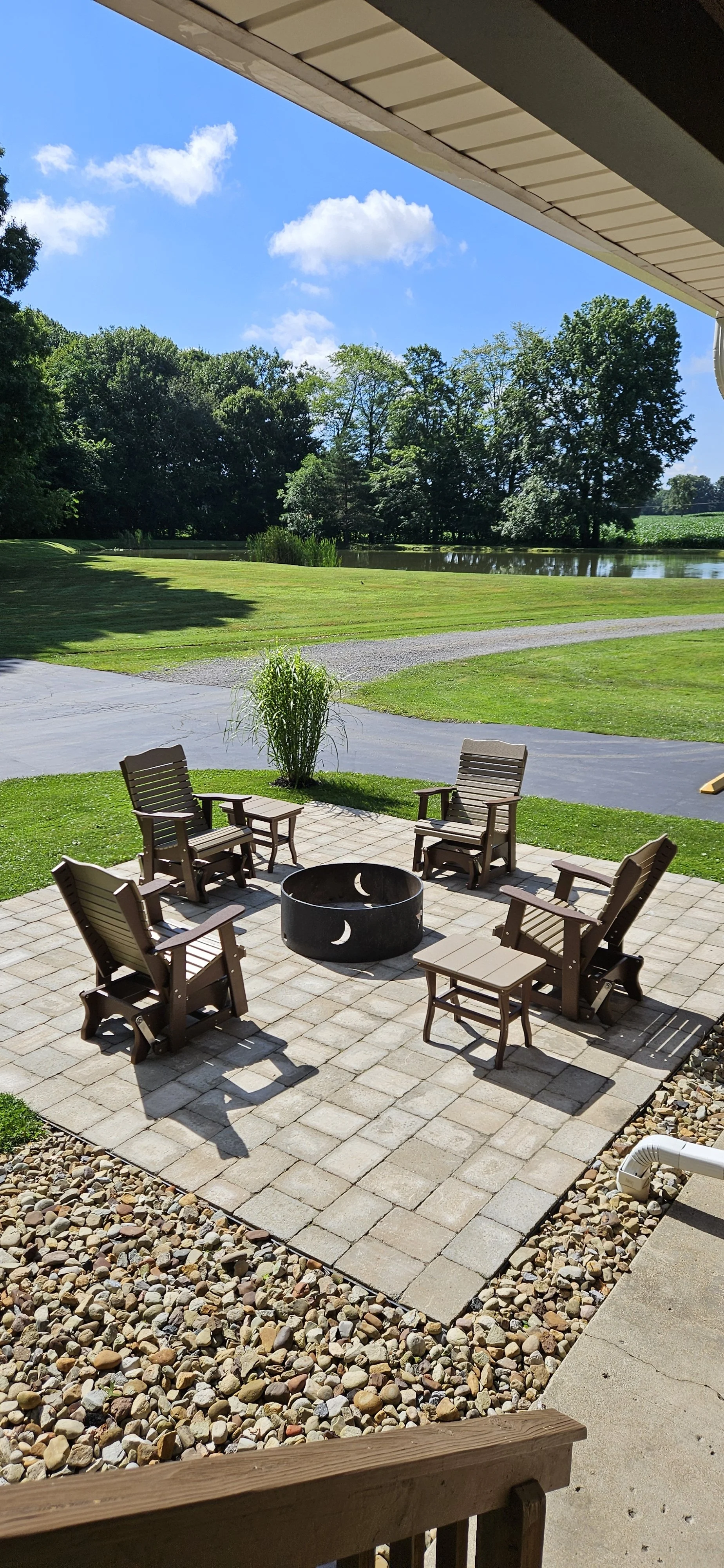 Patio with six wooden chairs and a fire pit, overlooking a grassy yard with a pond and trees under a blue sky with clouds.