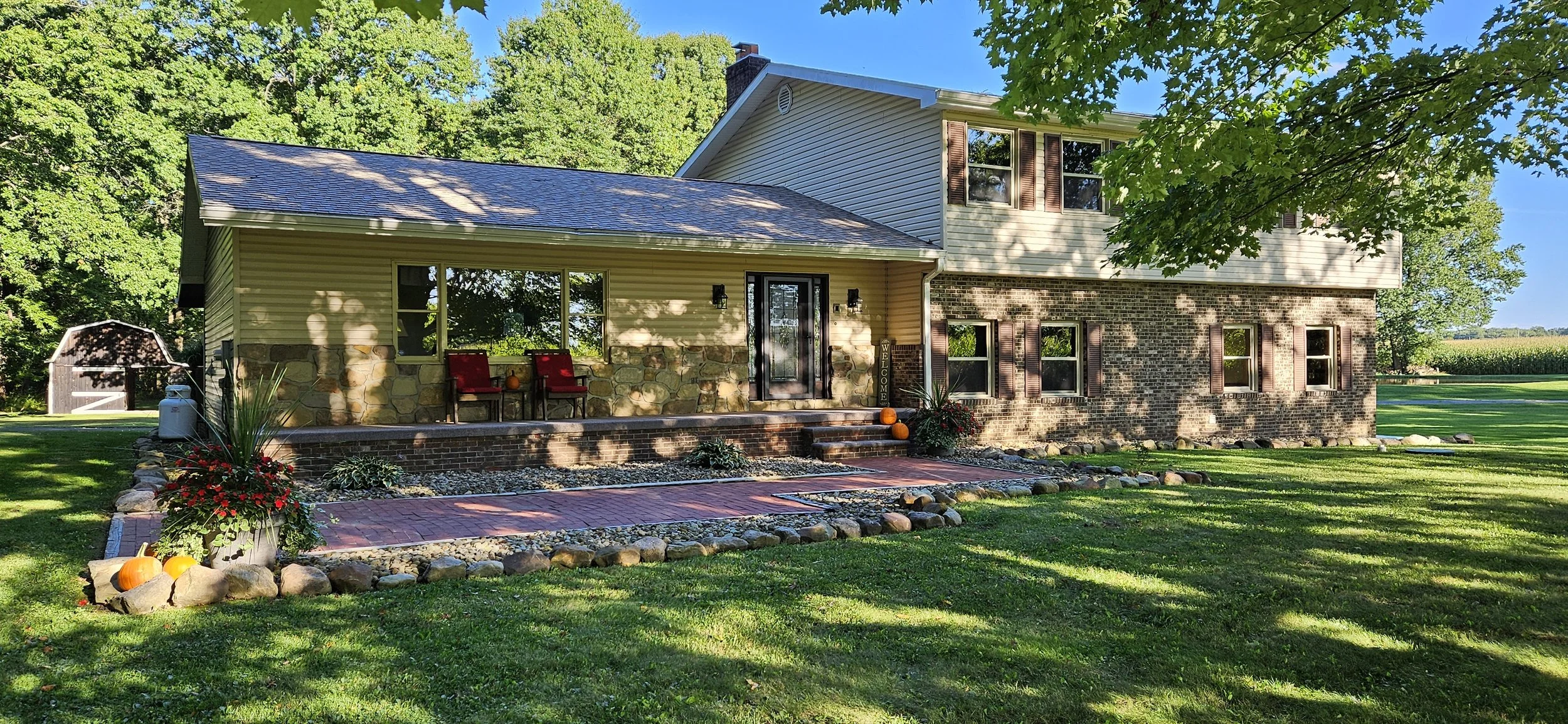 Front view of a two-story house with a stone and brick exterior, green lawn, and trees in the background. Decorated with pumpkins, potted plants, and chairs on the porch.