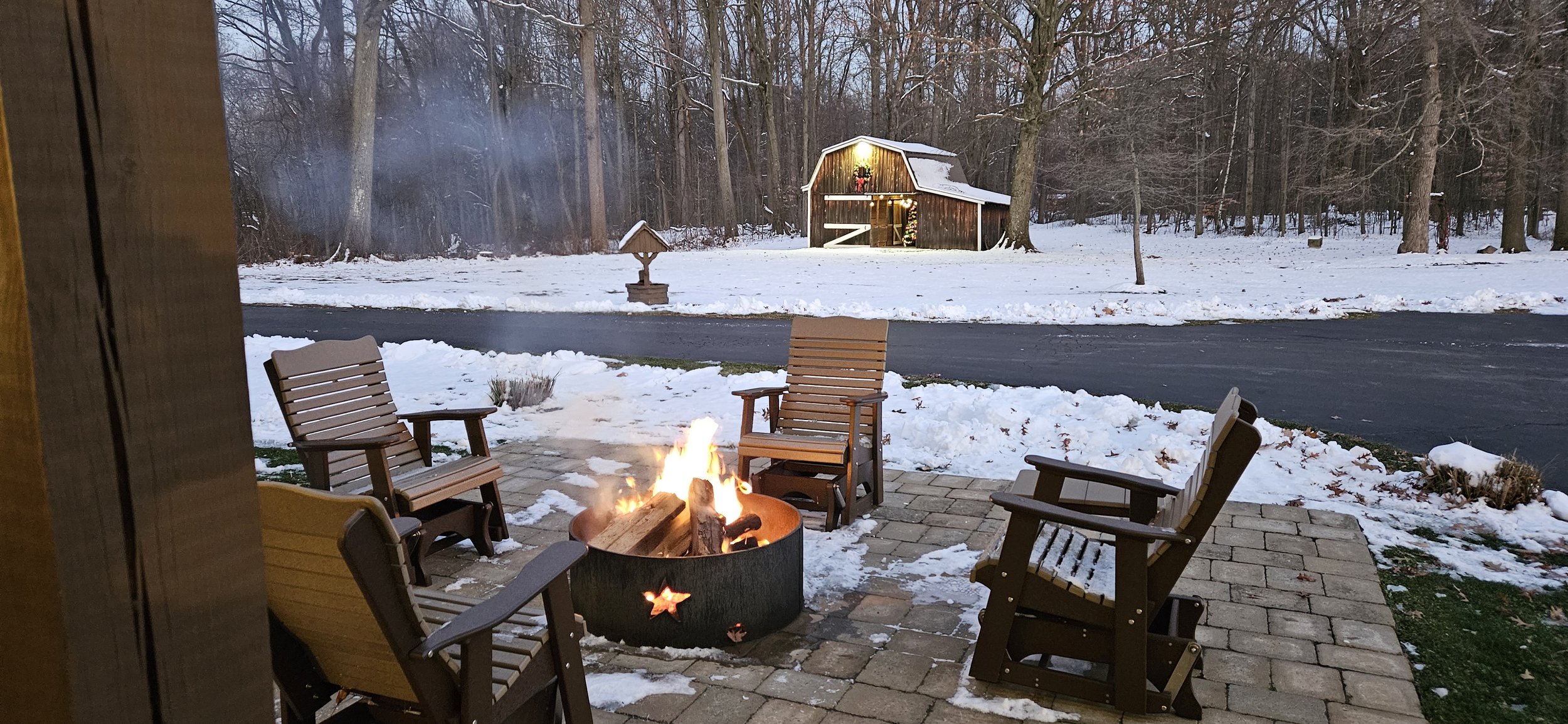 Backyard patio with six wooden chairs surrounding a lit fire pit on a brick surface, snow-covered ground and road beyond, and a rustic barn decorated with holiday lights in the distance.