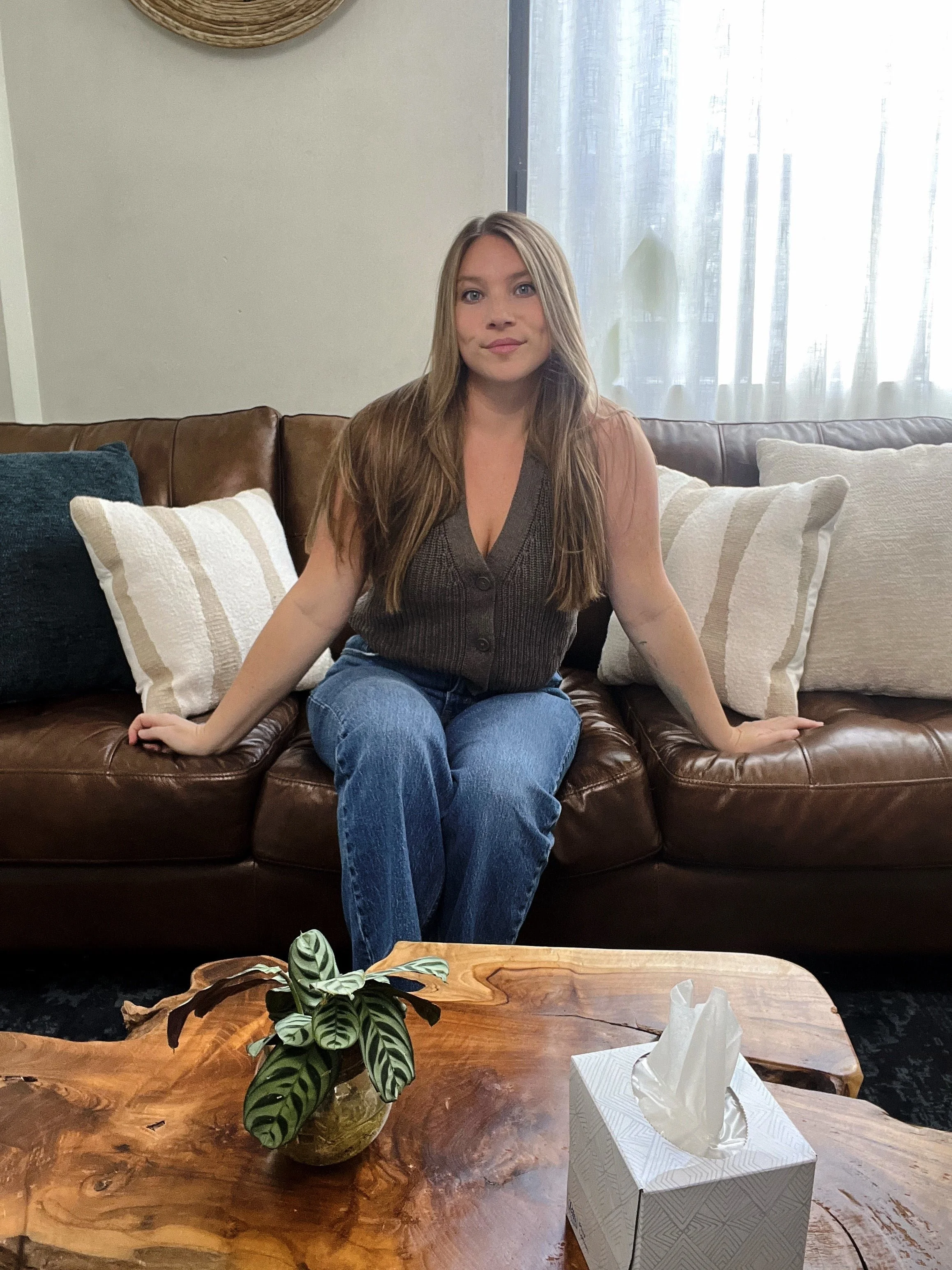 A young woman with long light brown hair sitting on a brown leather couch, with white and dark blue cushions, in a room with a window with sheer white curtains. In the foreground, a wooden table holds a potted plant with green patterned leaves and a box of tissues.