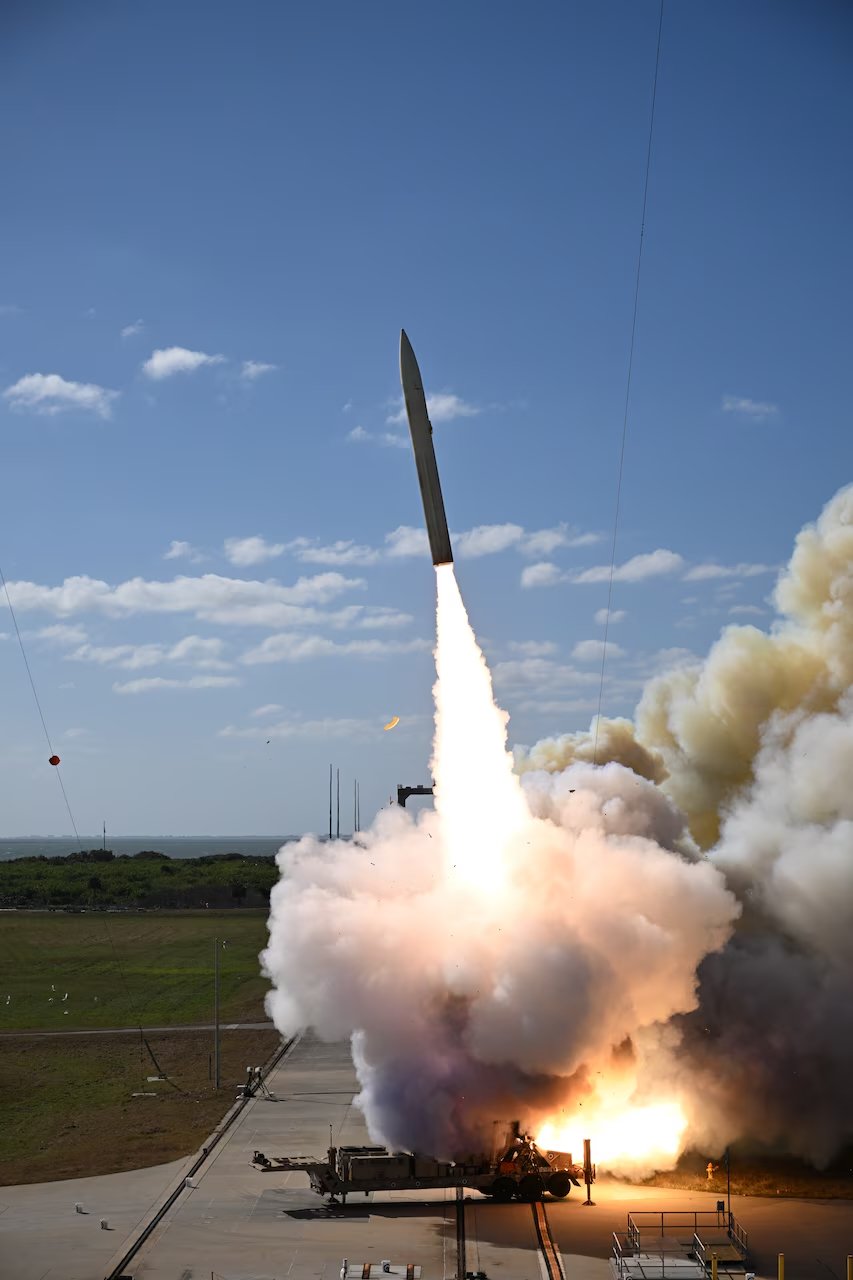 A missile being launched from a military vehicle on a flat open field with smoke and flames visible at the launch site. The missile is ascending into the sky during daytime with a partly cloudy sky in the background.