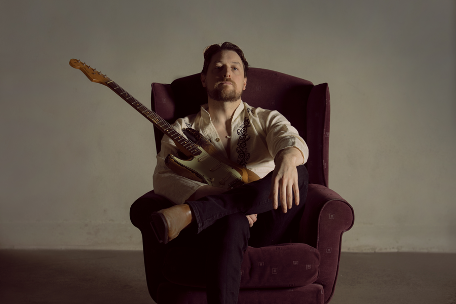 Brett Wood — Australian blues guitarist seated in an armchair holding an electric guitar against a neutral background