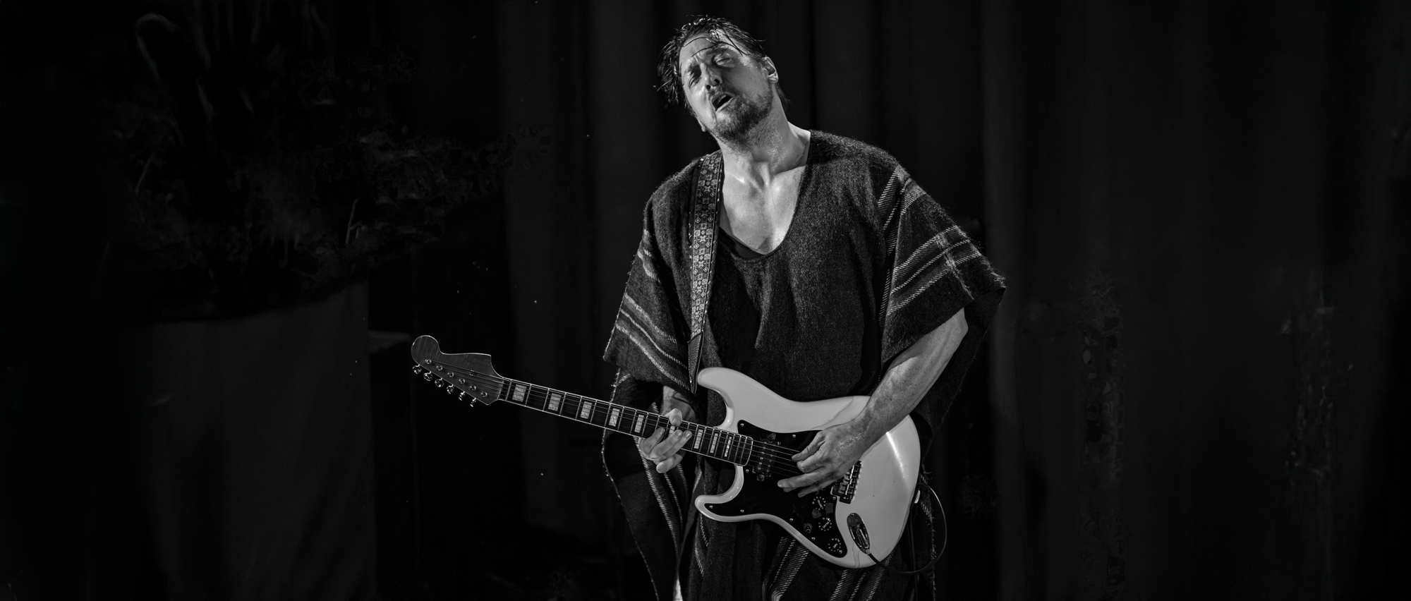A man playing an electric guitar on stage with a passionate expression, wearing a poncho and dark clothes, in a black and white photograph.