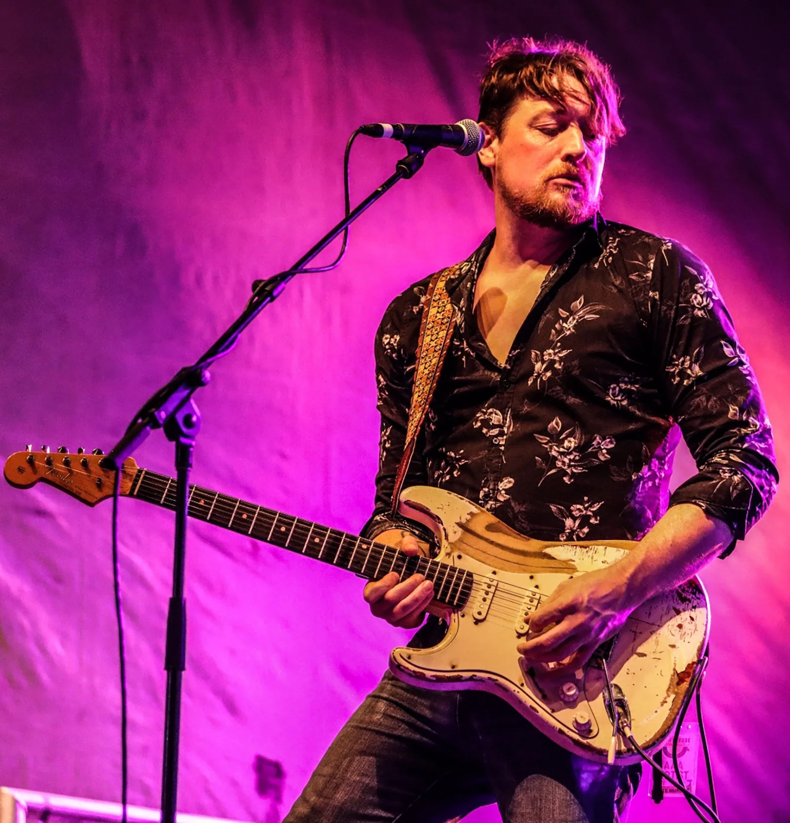 Brett Wood — Australian blues guitarist performing on stage under pink and purple lighting wearing a black floral shirt with a distressed electric guitar