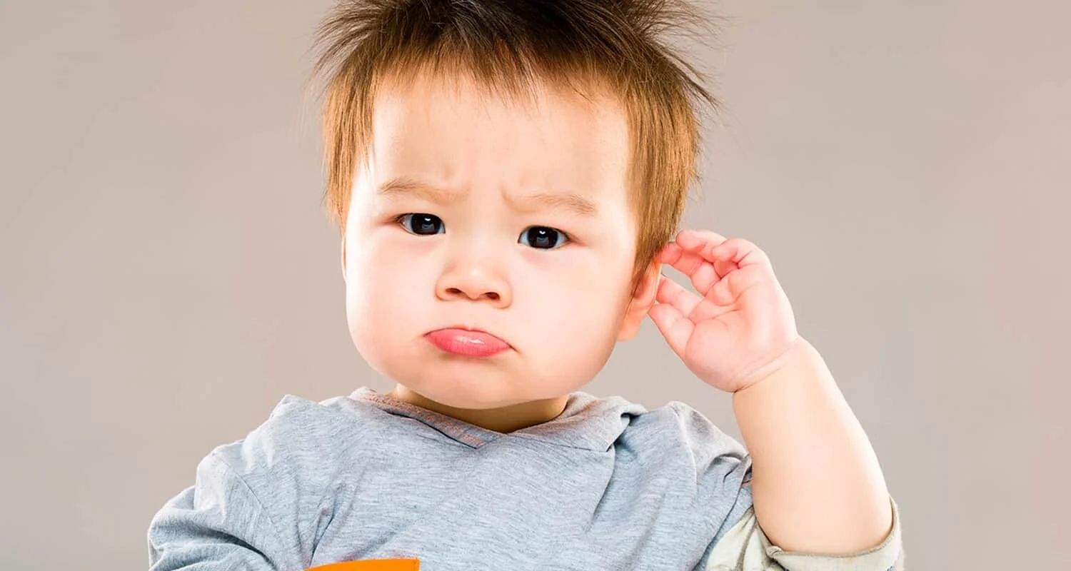 A young boy with brown hair and a puzzled expression holding his hand to his ear, suggesting he is listening.