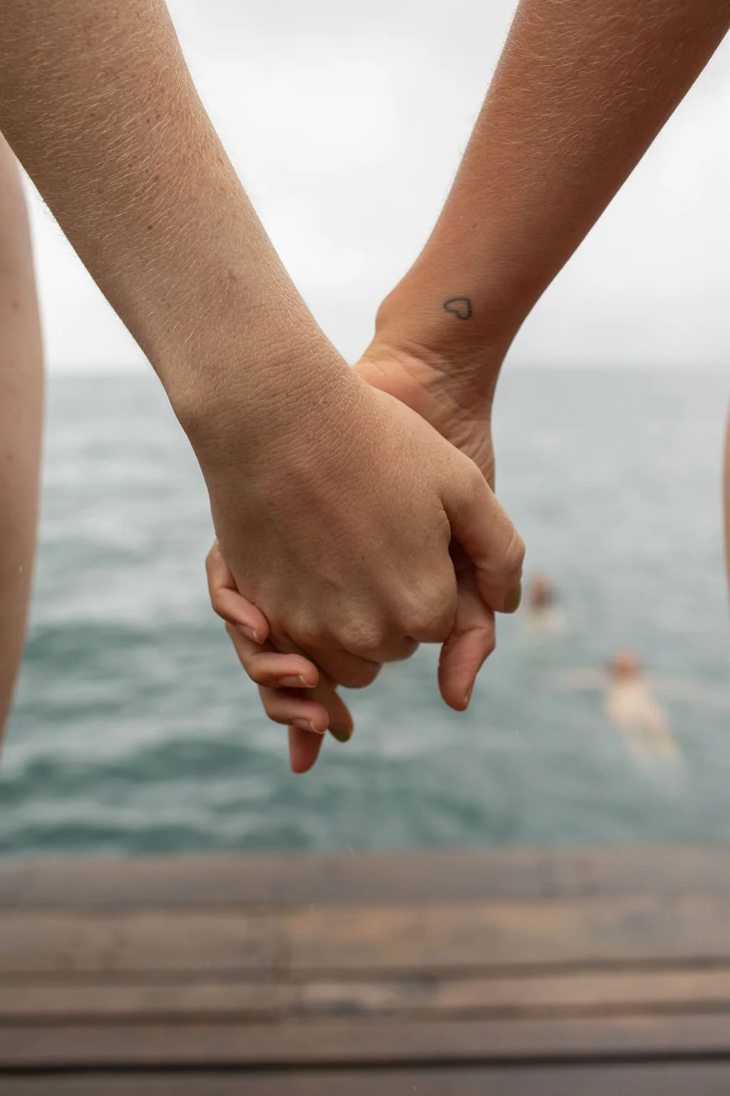 Two people holding hands over a body of water, with one person having a small black heart tattoo on their wrist.