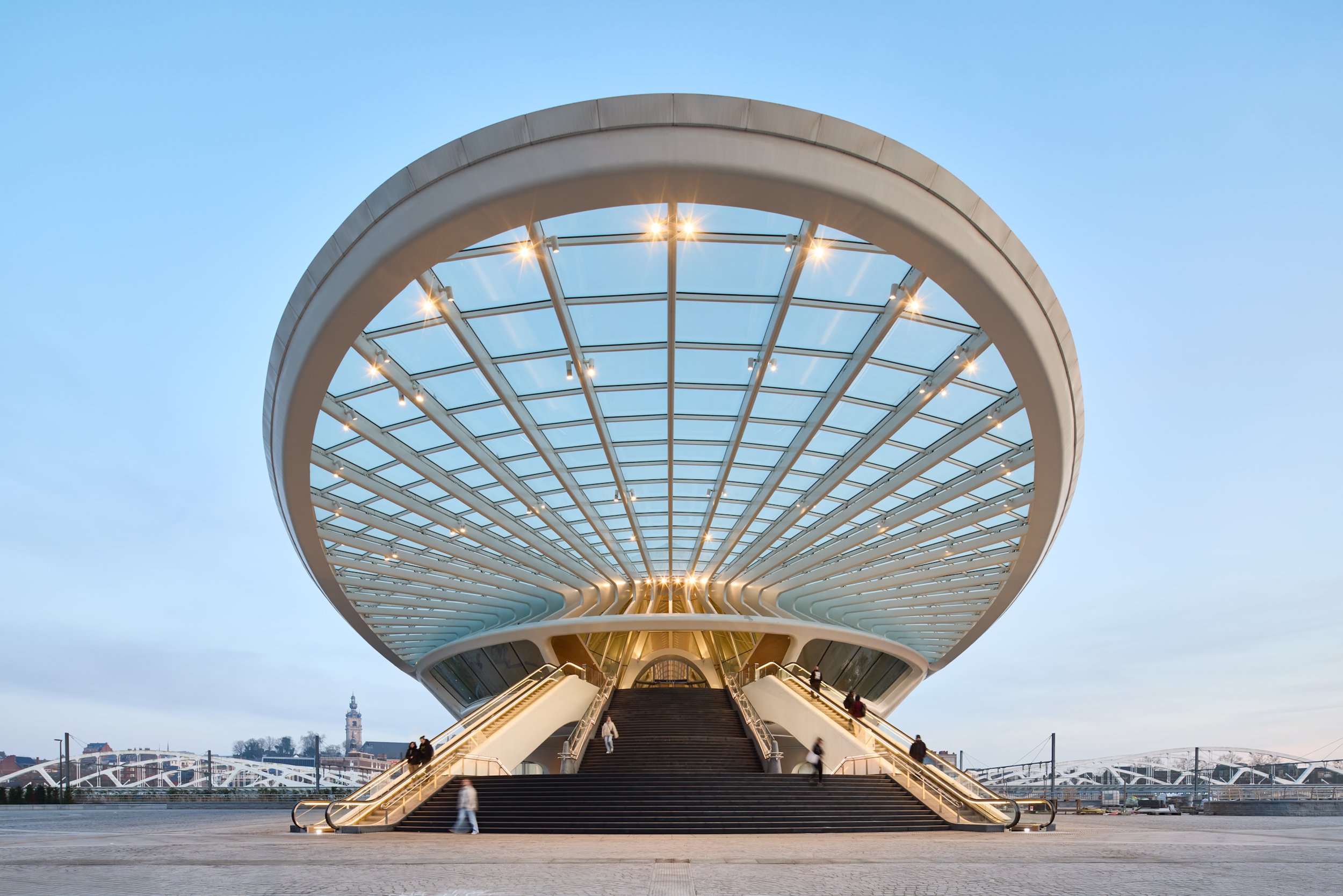 Modern building with a futuristic design, featuring a large, circular glass roof and wide stairs leading up to the entrance, with a few people walking.
