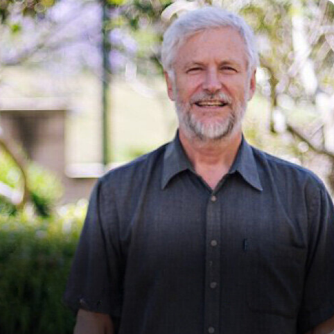 Man with white hair and beard wearing a dark shirt smiles outdoors with blurred greenery in the background.