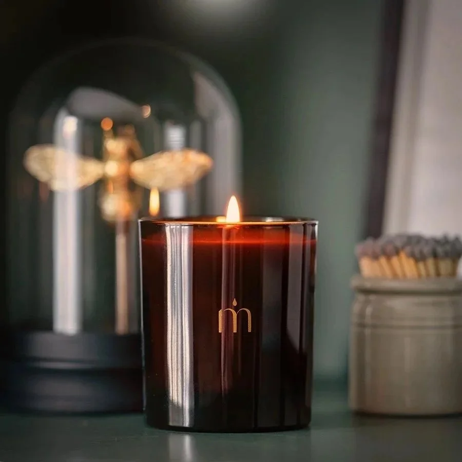A lit black candle with a gold 'm' on it, placed on a green surface, with a blurred decorative glass jar and a jar of matches or sticks in the background.