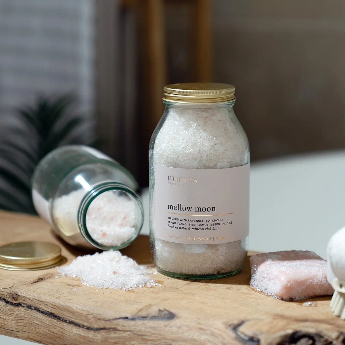A large glass jar labeled 'mellow moon' containing bath salts, with some salts spilled on a wooden surface, an open jar of salts lying on its side, and a pink bath soap bar with some foam nearby.