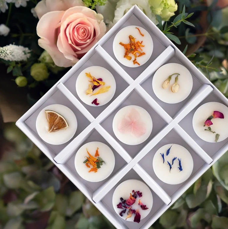 A box of nine decorative botanical wax melts with floral and fruit embellishments, surrounded by pink and white roses and greenery.