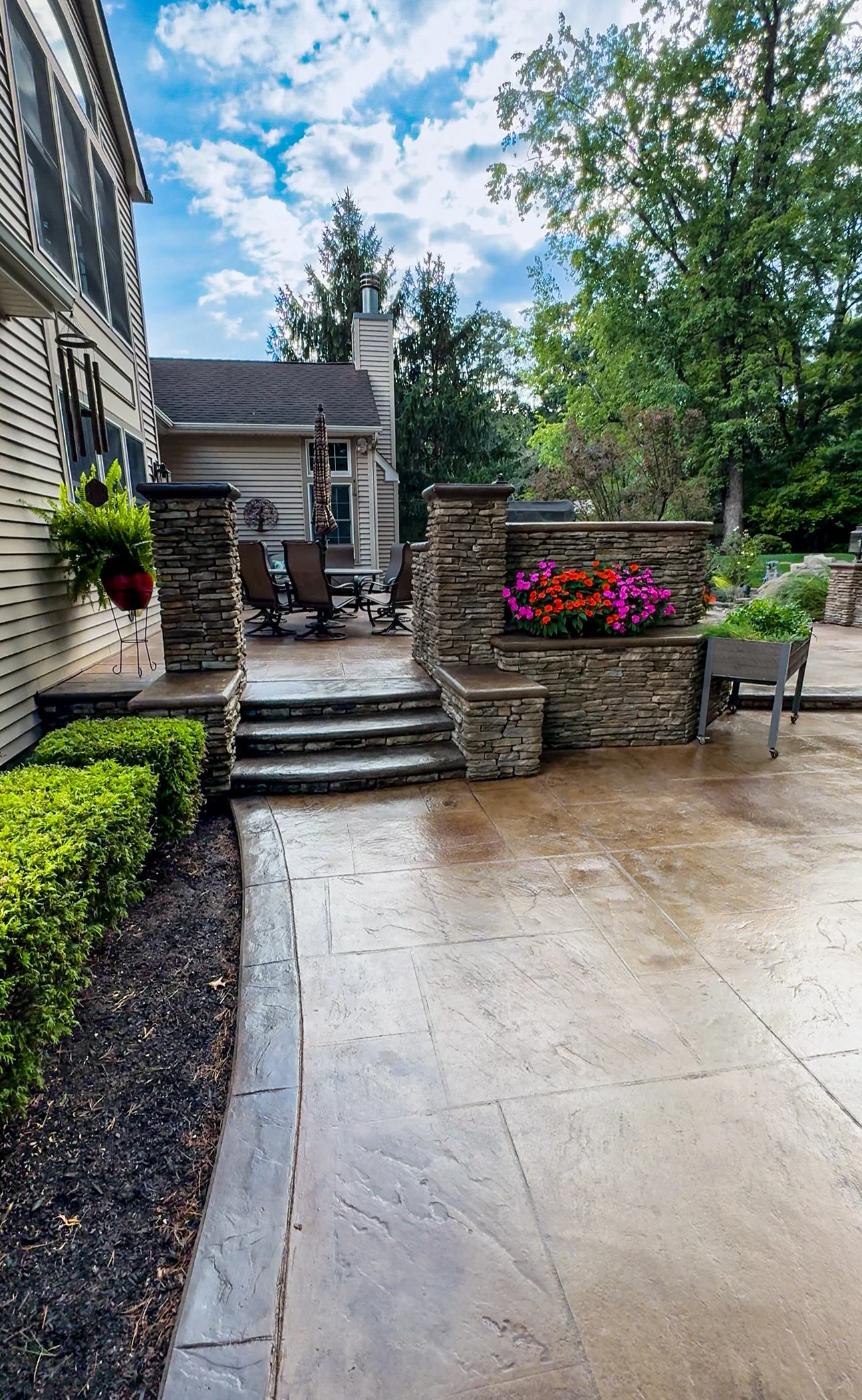 A backyard patio area with stone steps leading up to a seating area with a table and chairs. There are colorful flowers in planters, lush green bushes, and trees with a partly cloudy sky overhead.