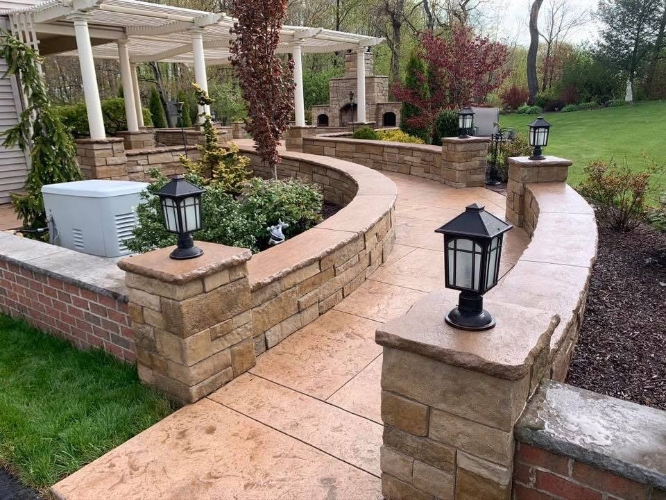 A curved stone patio with wall and lanterns, garden area with plants and trees, and a white pergola in the background, in a backyard setting.