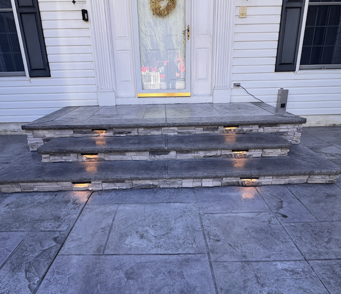 Stone steps leading up to a white front door with a holiday wreath, surrounded by white siding and black shutters. The steps have built-in lights illuminating the edges.