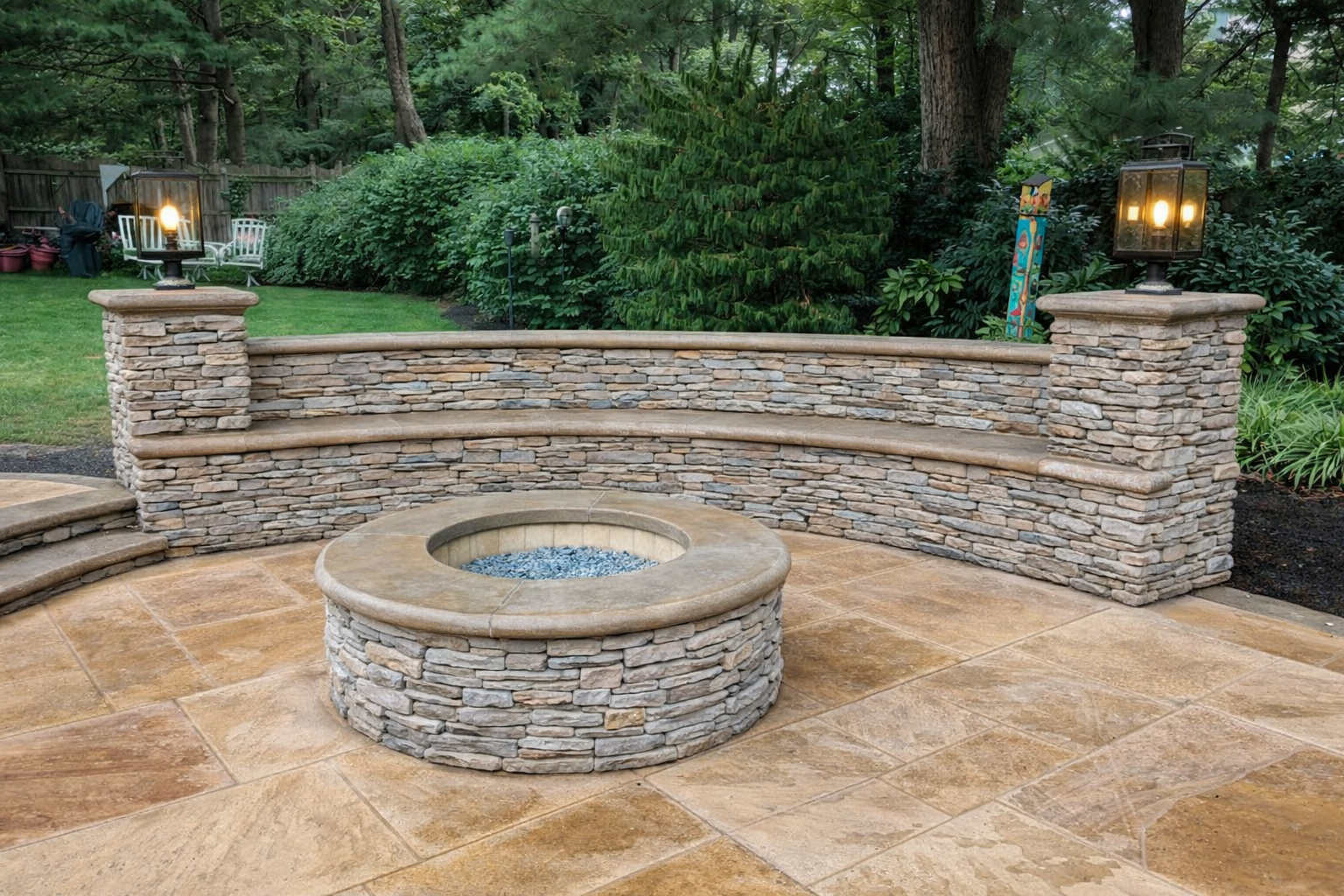 A backyard with a stone fire pit, surrounding flagstones, and a stone retaining wall topped with a concrete cap. Lantern-style outdoor lights are on each end of the wall, and trees and shrubs are in the background.