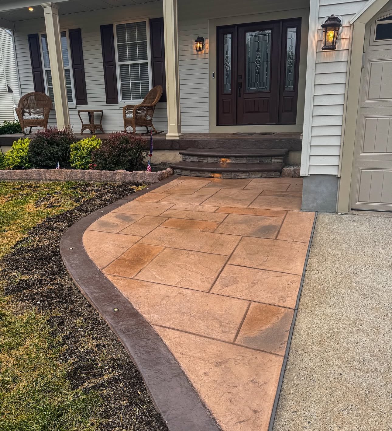 The image shows the front porch of a house with a new stamped concrete walkway, landscaped garden beds, and outdoor seating area. The house has a dark wooden front door, two lantern-style wall lights, and decorative shutters.