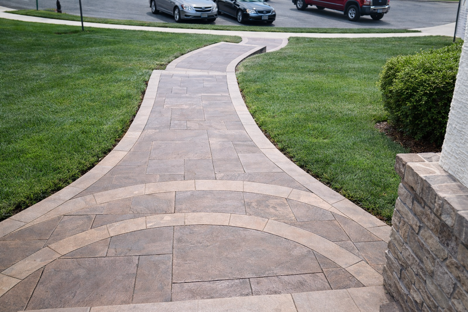 A paved walkway with a curved design running through a well-maintained lawn, with parked cars in the background and a stone structure on the right.