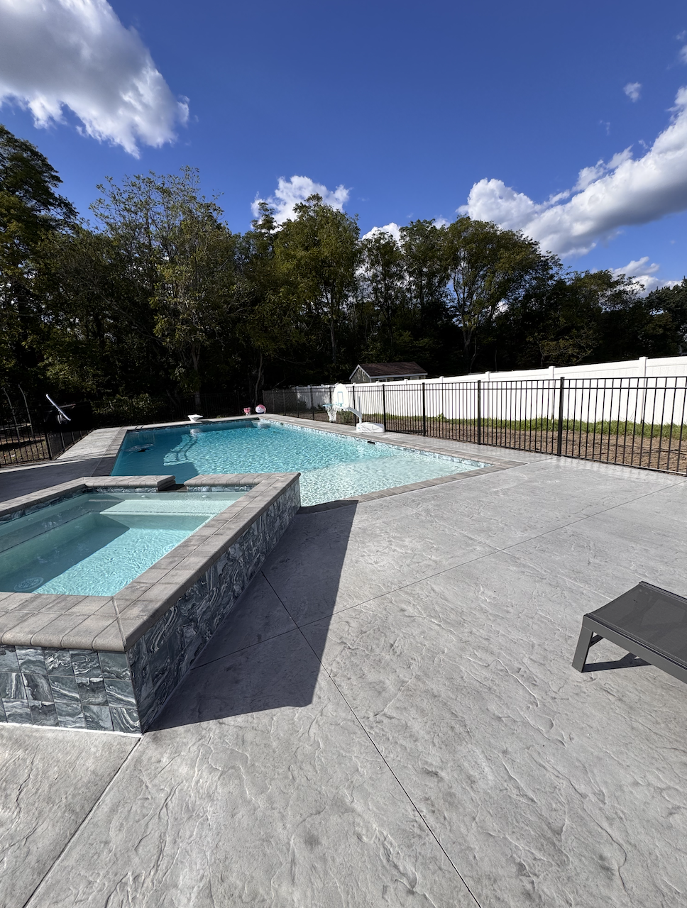 View of a backyard with a concrete patio, swimming pool, hot tub, and a black fence, surrounded by trees under a cloudy blue sky.