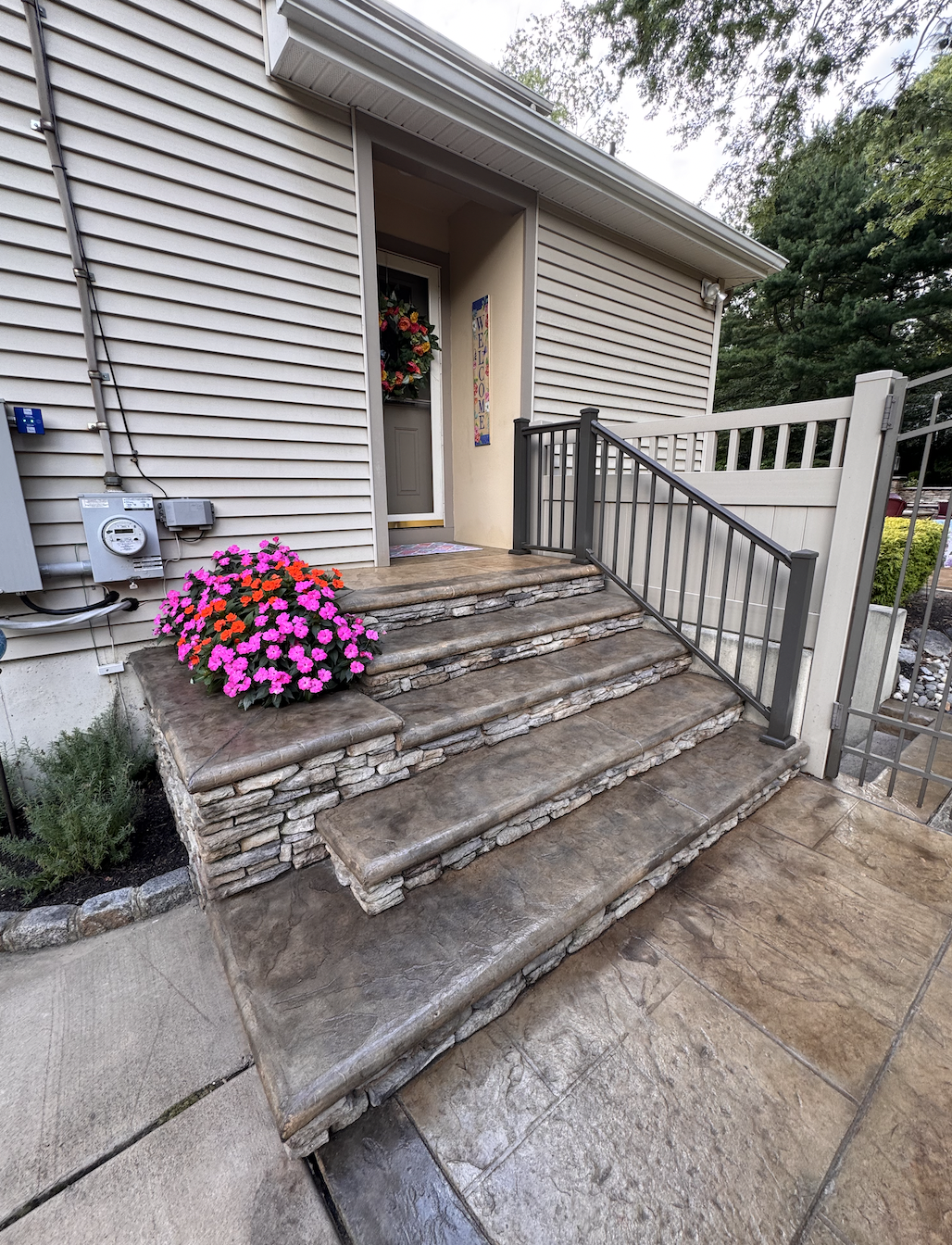 Front porch with stone steps, railing, potted pink and orange flowers, welcome sign, and a door with a flower wreath.