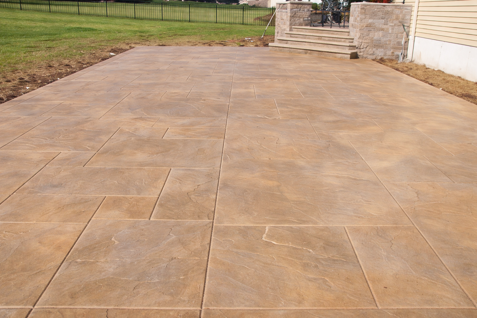 A newly installed beige concrete patio with large tiles, surrounded by a grassy yard and a house with steps leading to a porch.