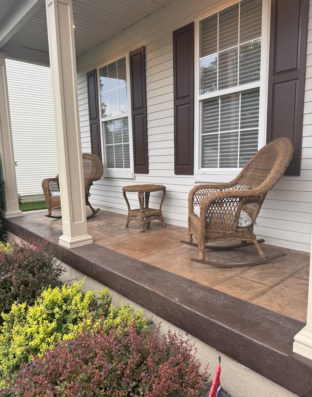 Front porch with two wicker rocking chairs, a small wicker table, and bushes with pink and yellow flowers in front of a house with white siding and two large windows with brown shutters.