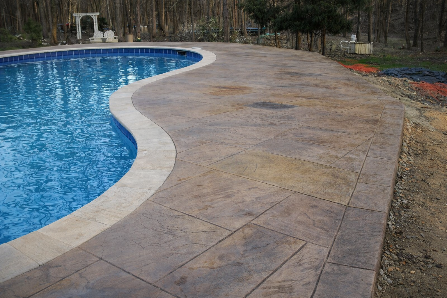 Close-up view of a newly constructed swimming pool with a curved edge, surrounded by a textured concrete deck and a wooded background.