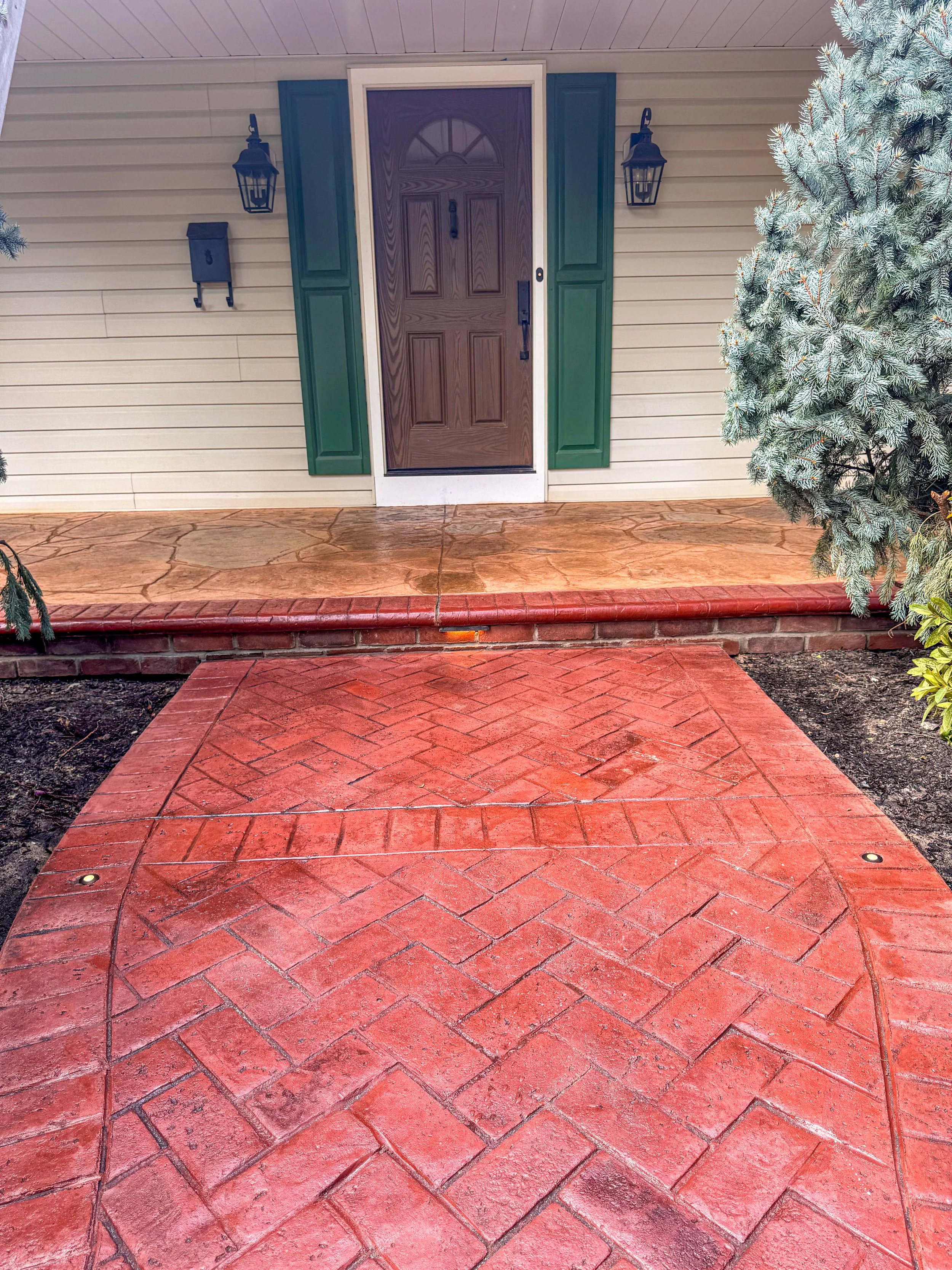 Front porch with a wooden door, green shutters, and outdoor lighting fixtures, with a red brick walkway and a shrub on the right side.