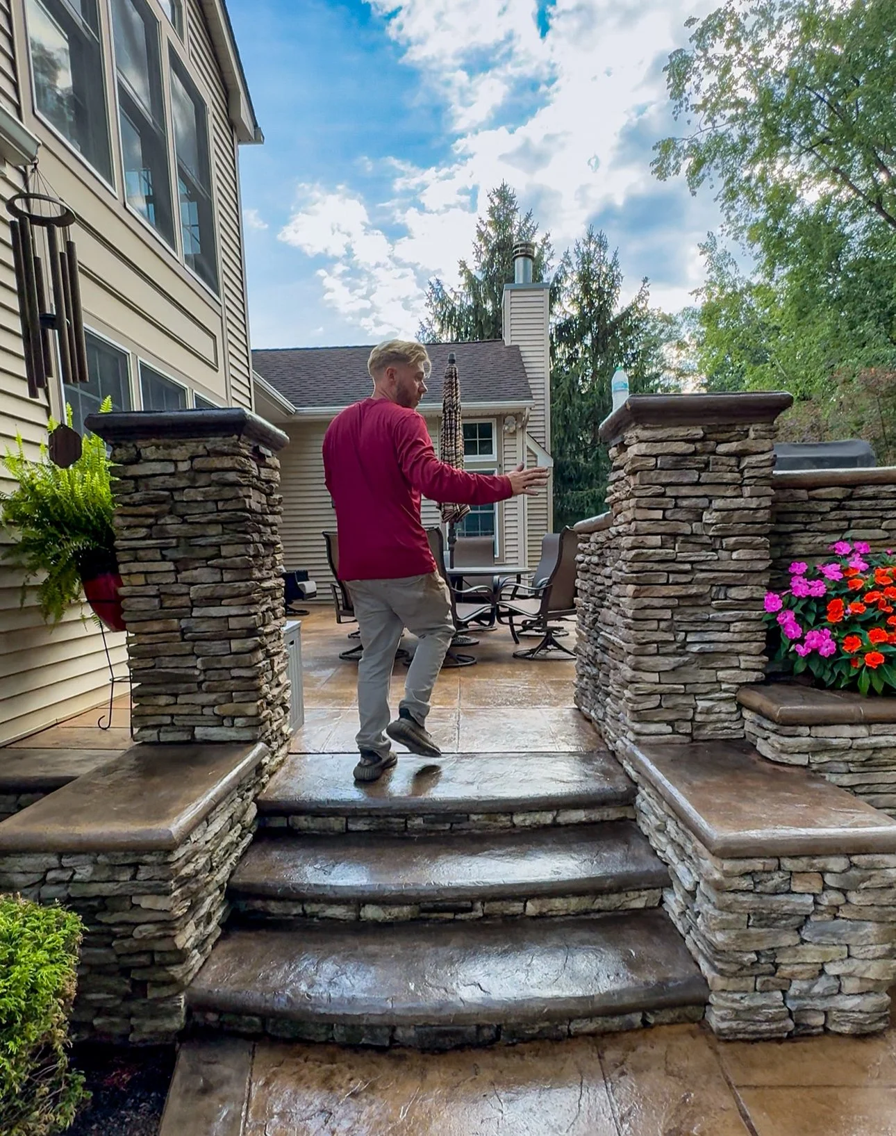 A man in a red shirt and light-colored pants standing on stone stairs in a backyard patio, with trees, flowers, house, and sky in the background.