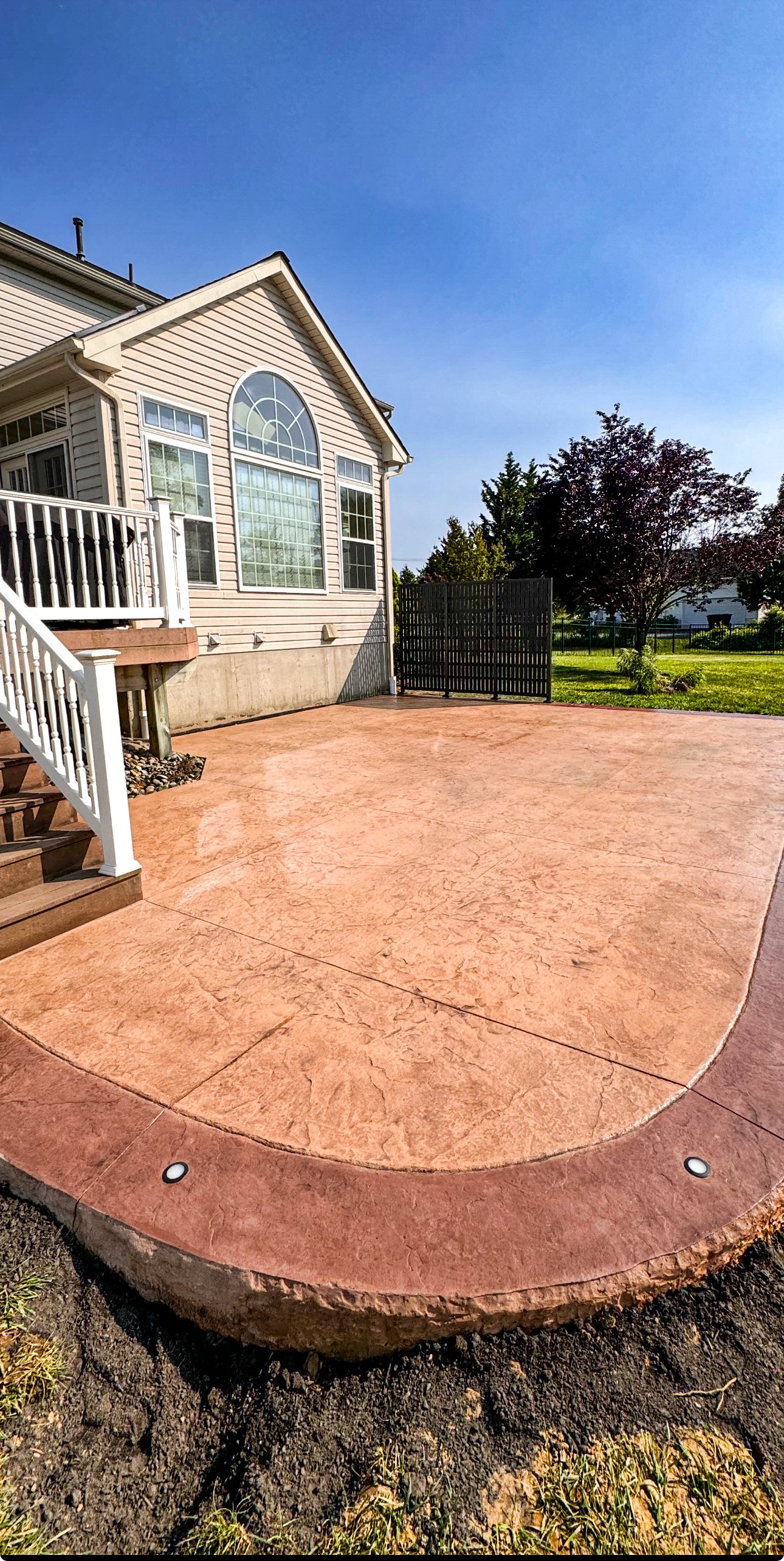Backyard with a stamped concrete patio, beige house with large arched window, white railing staircase, and trees in the background under a blue sky.