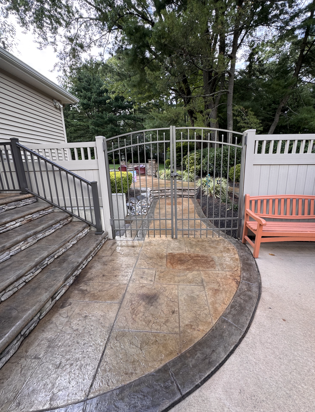 Backyard patio with stone steps leading to a gated area, garden, and trees in the background, with a pink bench on the right side.