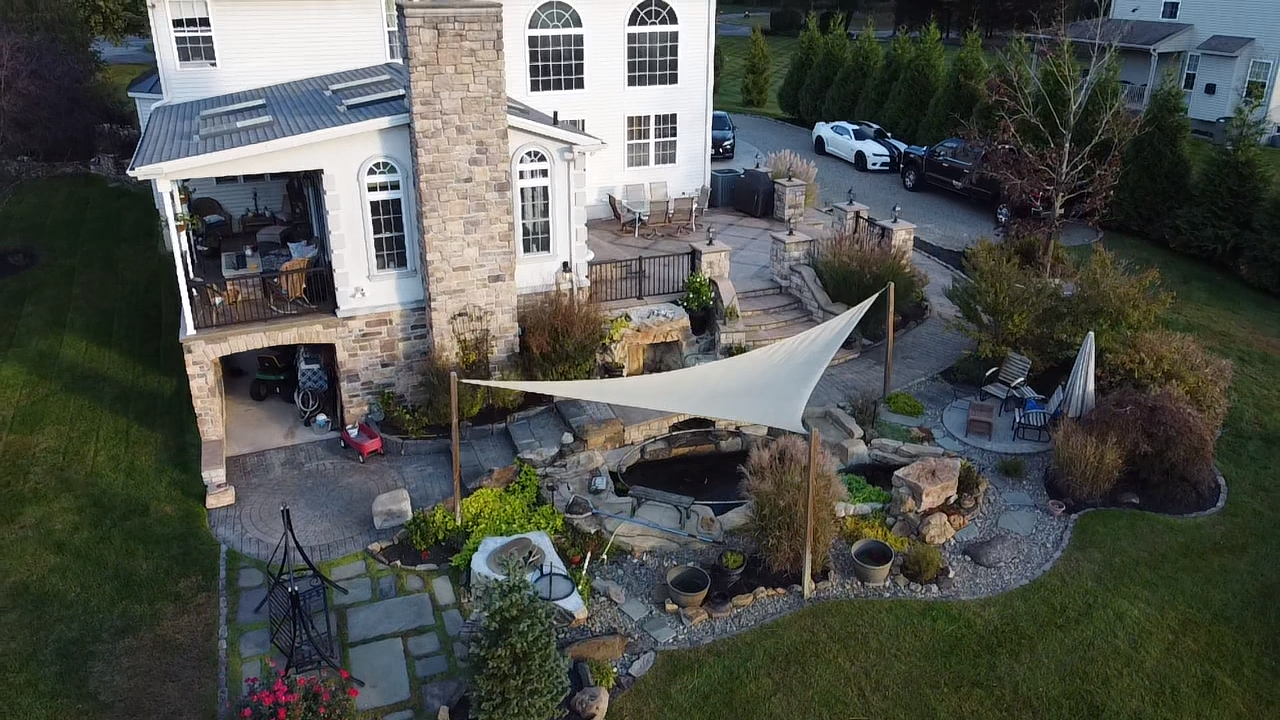 Aerial view of a backyard patio and garden area behind a large white house with multiple levels, a stone chimney, and a garage. The patio features a pool with a shade sail, outdoor seating, and landscaped plants and trees.
