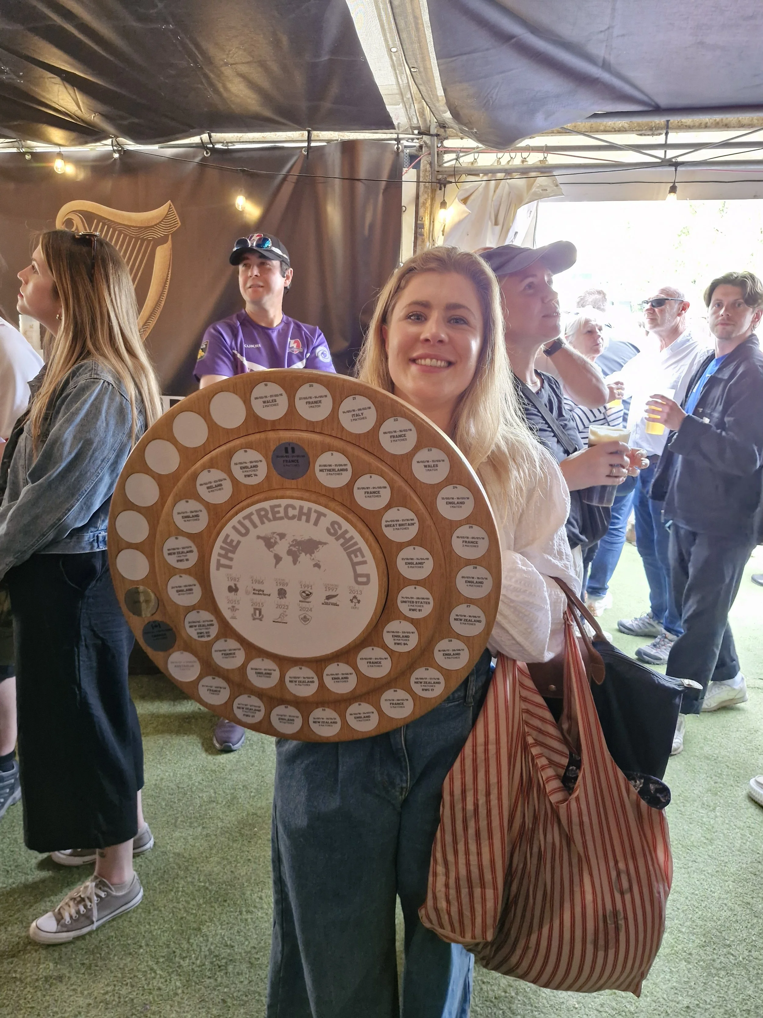 A woman holding a circular wooden sign that reads 'The Utrecht Shield' with a world map and years, at a crowded outdoor event. Other people are seen in the background under a tent.