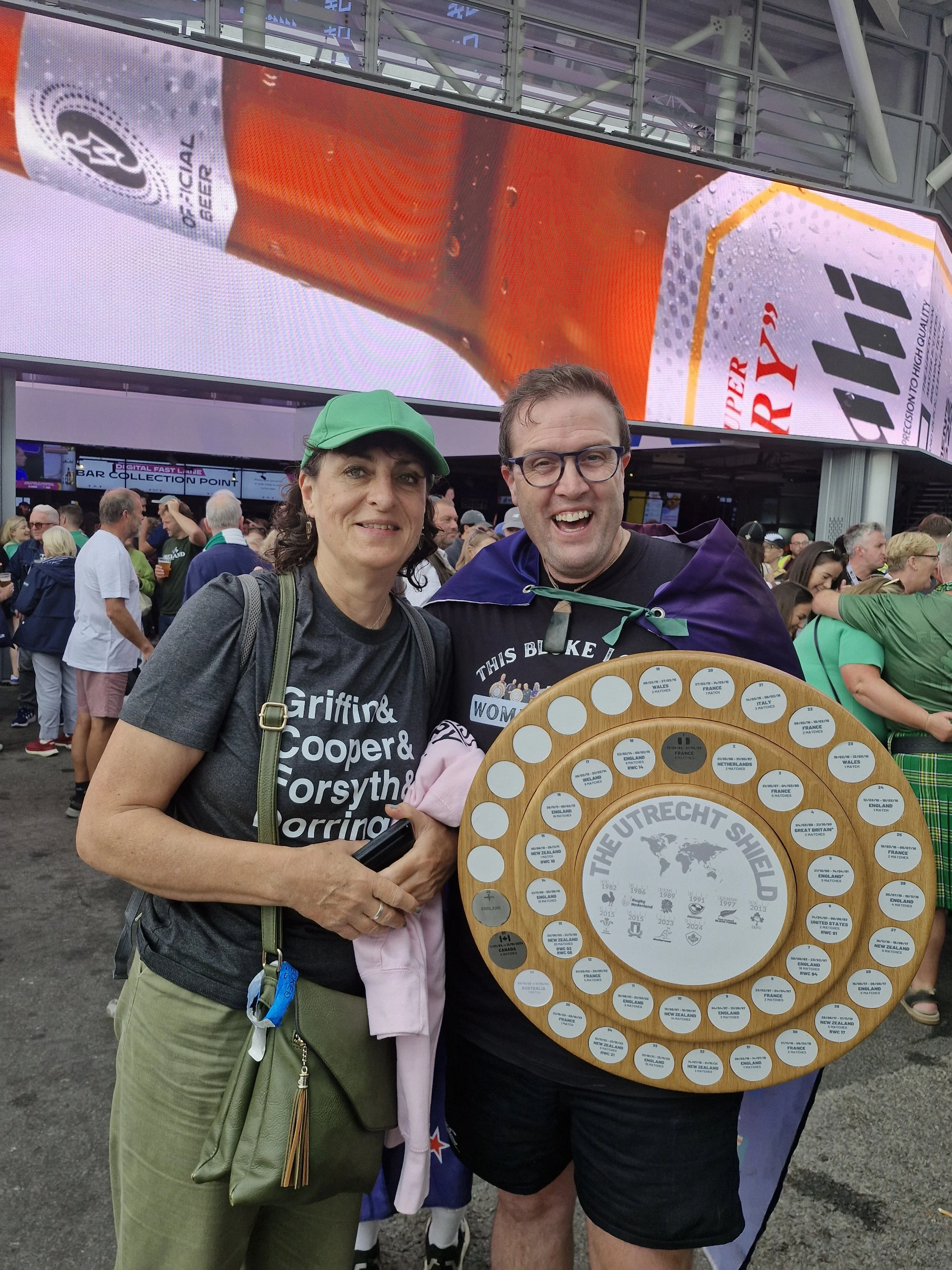 Two people standing together at a crowded event. The person on the left is wearing a gray T-shirt with text, a green cap, and khaki pants, holding a purse and pink cloth. The person on the right is wearing glasses, a black T-shirt, with a purple cape