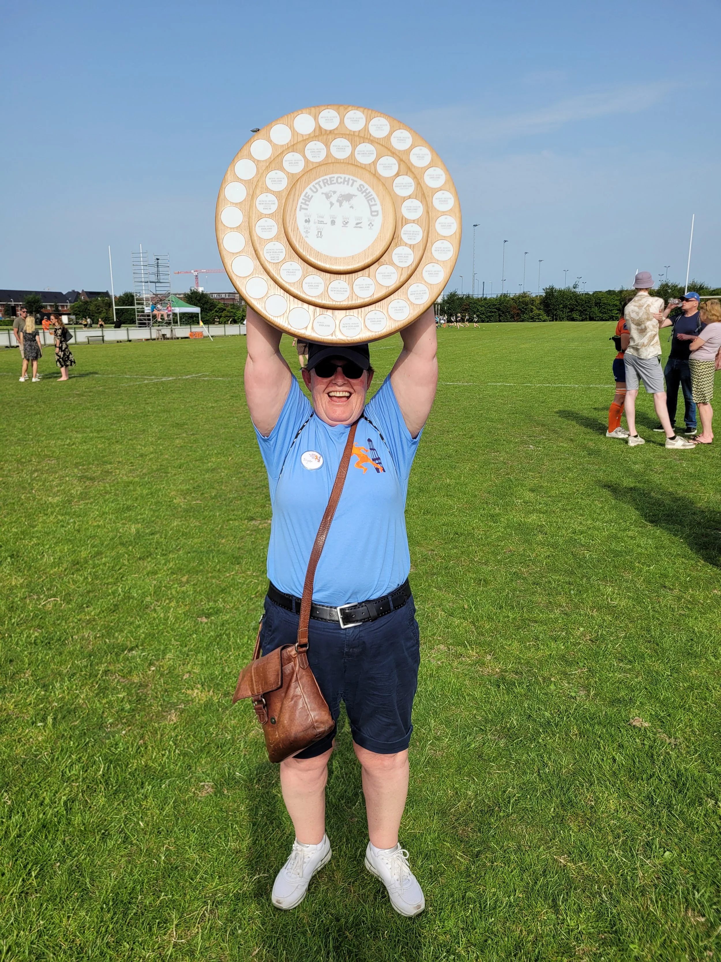 A person in a blue shirt and shorts, wearing sunglasses and a cap, smiling and holding a large circular award or shield above their head. They are standing on a grassy field with other people in the background, some standing and chatting, and others 