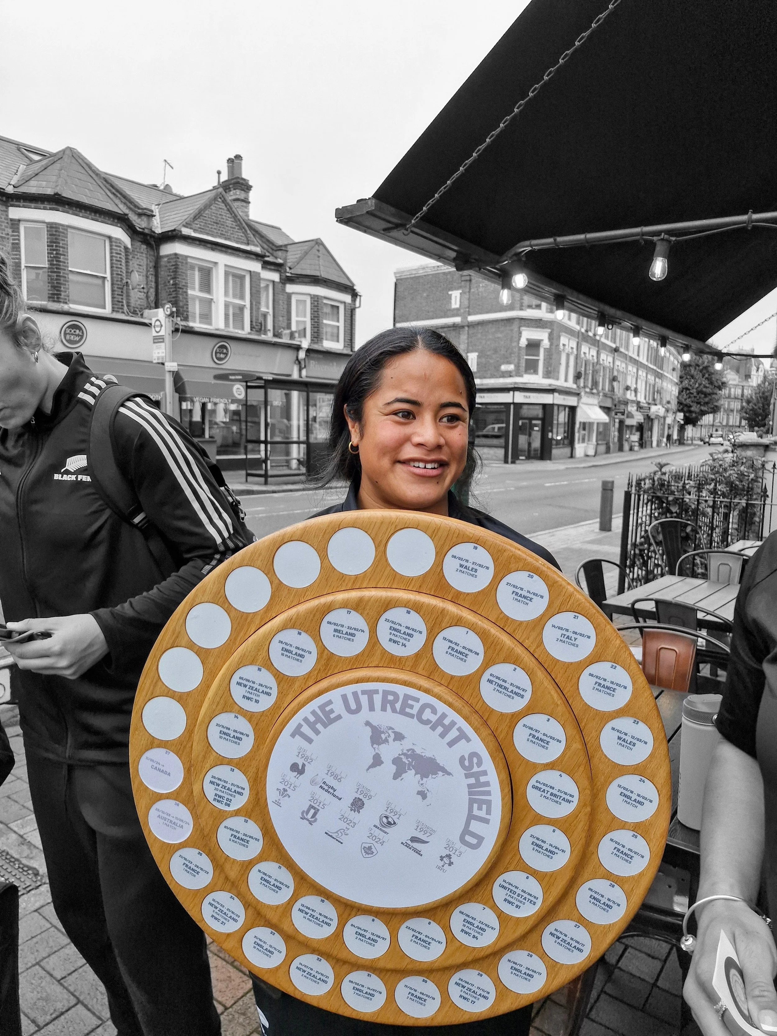 Young woman holding a circular wooden display board titled 'The Utrecht Shield' with various country flags and names, outdoors on a city street.