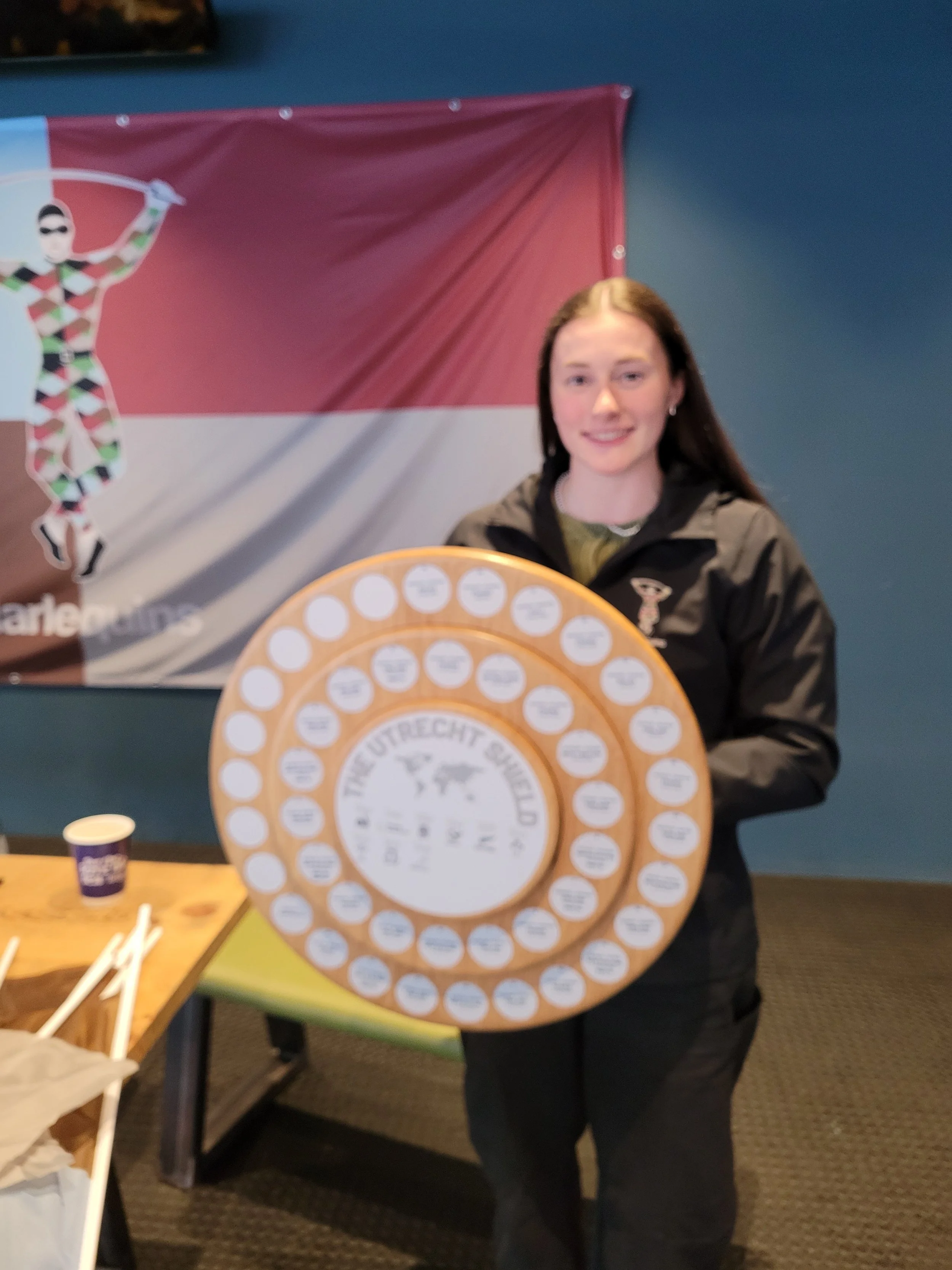 A woman holding a large circular cheese wheel with a logo that reads 'De Utrechtse Kaas'. She is indoors, standing in front of a colorful banner featuring a cartoon character.
